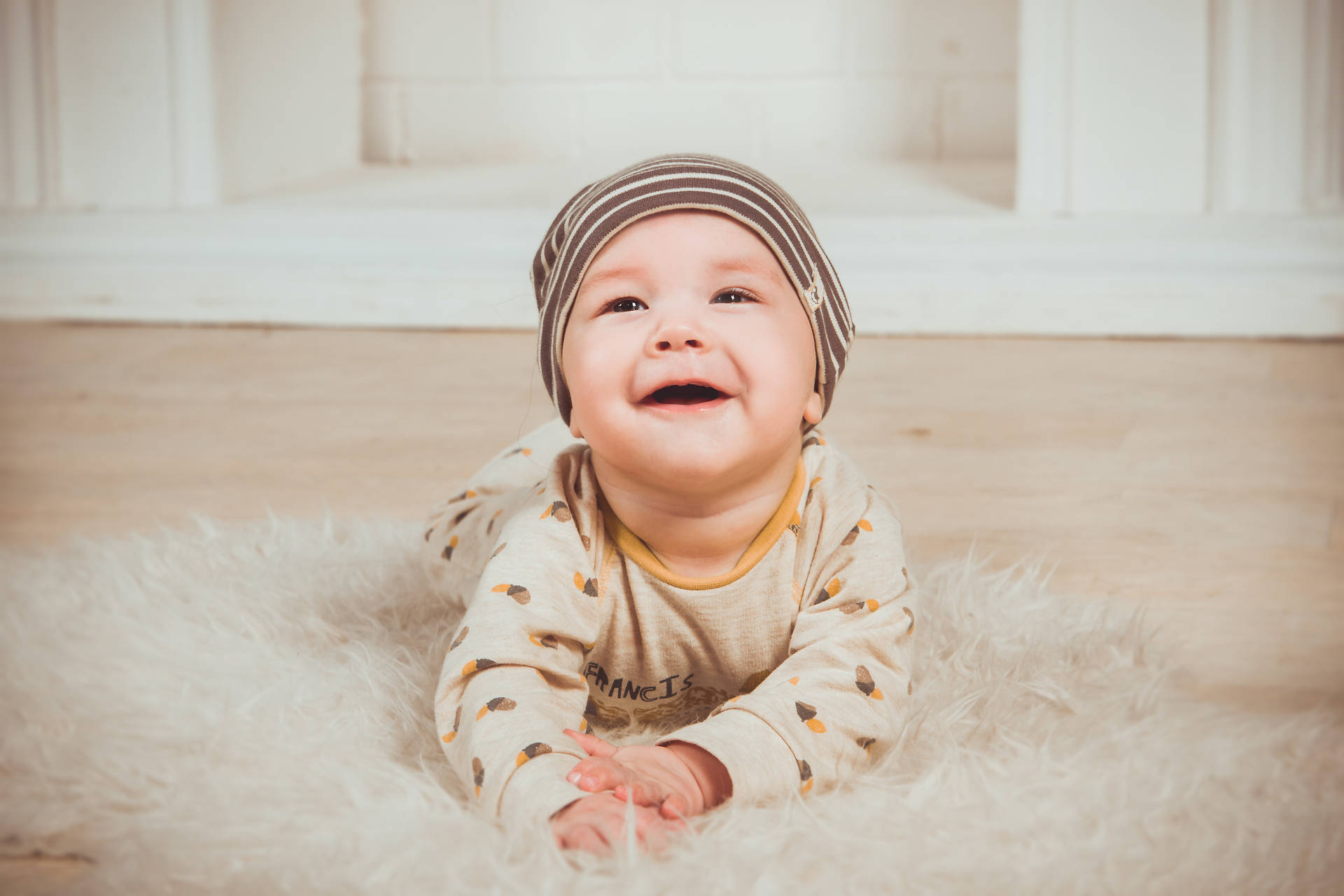 笑容燦爛的可愛嬰兒 壁紙 — 一個快樂的嬰兒戴著條紋帽，躺在室內柔軟的白色地毯上，開懷大笑。