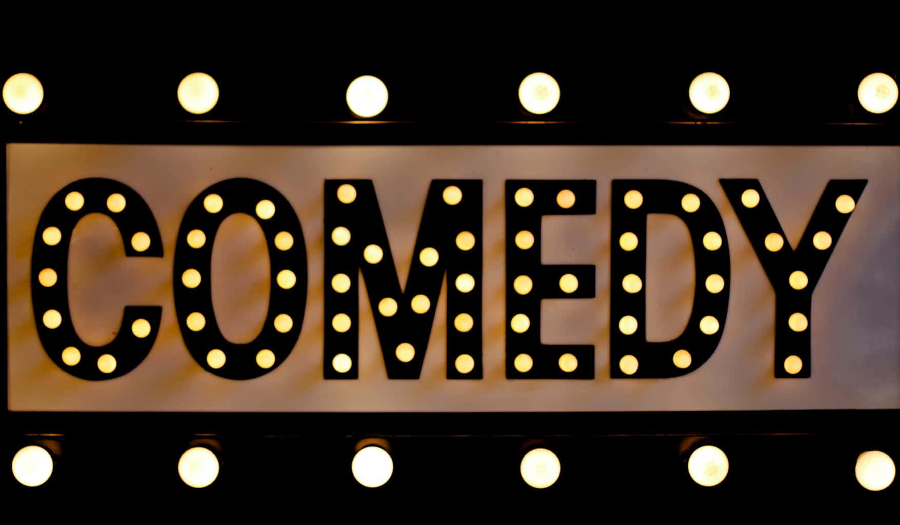 笑是最佳良藥 壁紙 — 一個發光的招牌，以粗體字母拼出COMEDY，周圍環繞著溫暖的圓形燈泡，背景為深色。