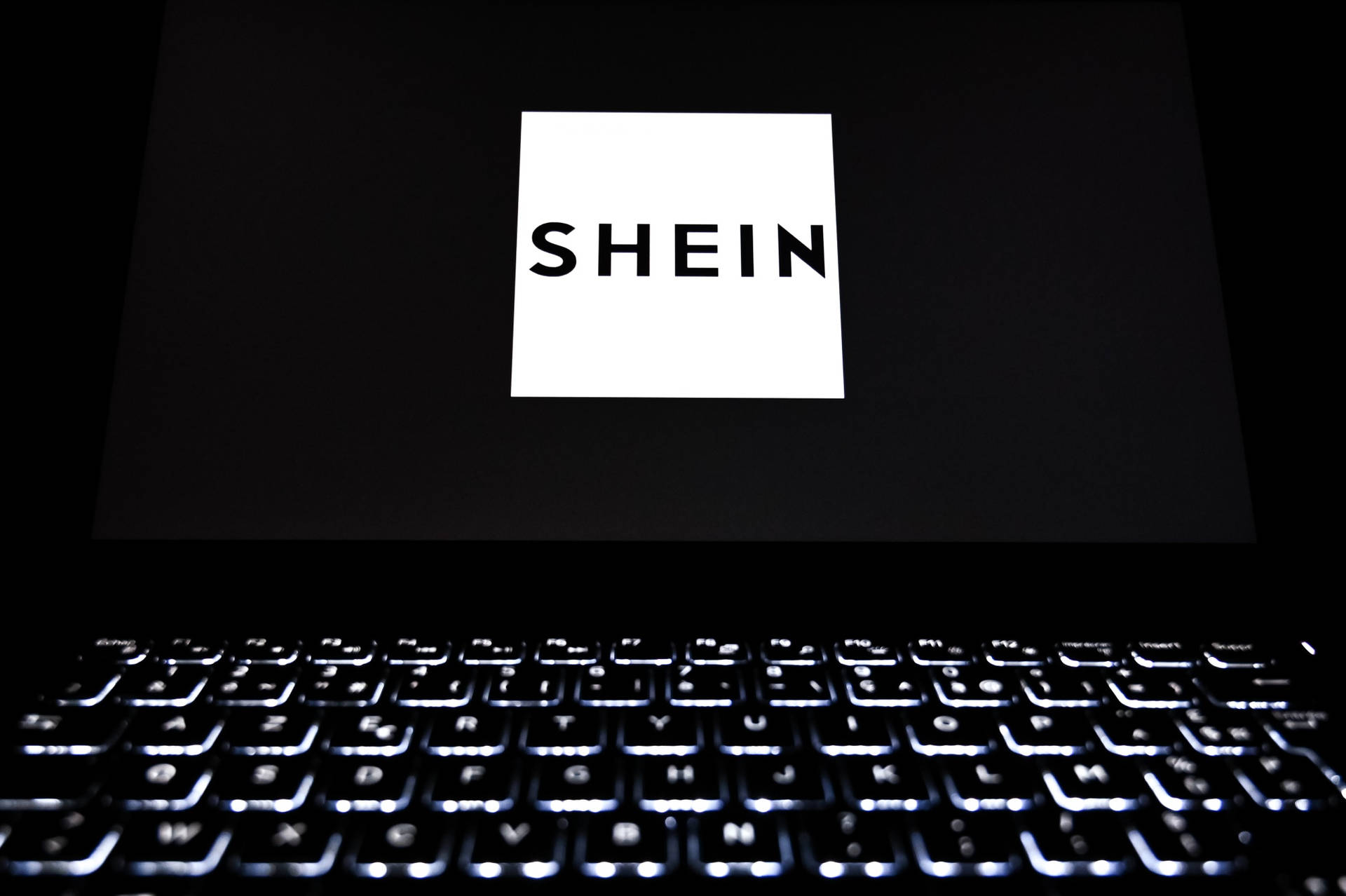 手提電腦屏幕顯示SHEIN標誌，鍵盤在黑暗房間中發光