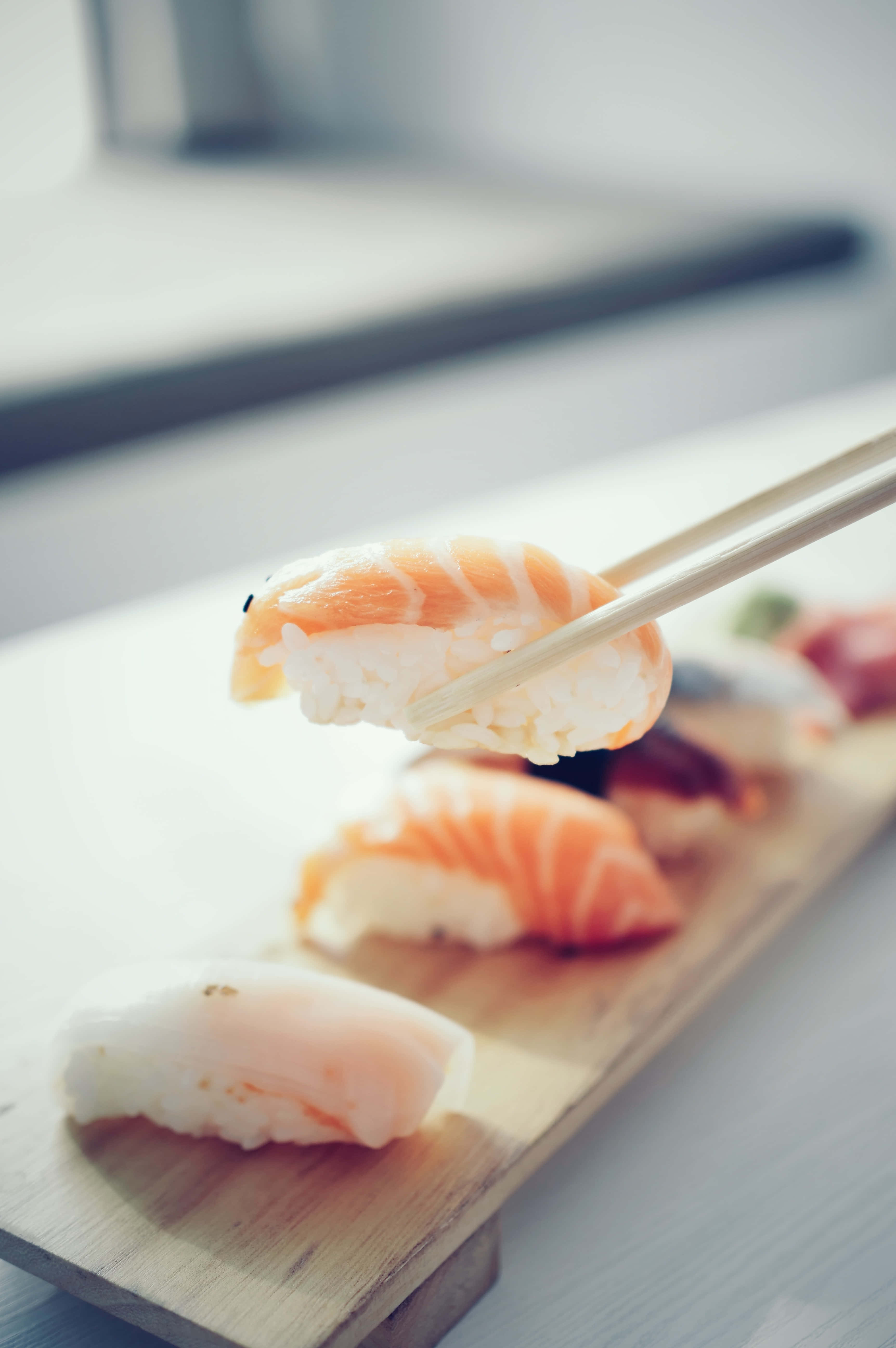 Sushi 桌布 壁紙 — 筷子從木盤上夾起一片三文魚握壽司的特寫，背景中其他壽司片模糊不清。