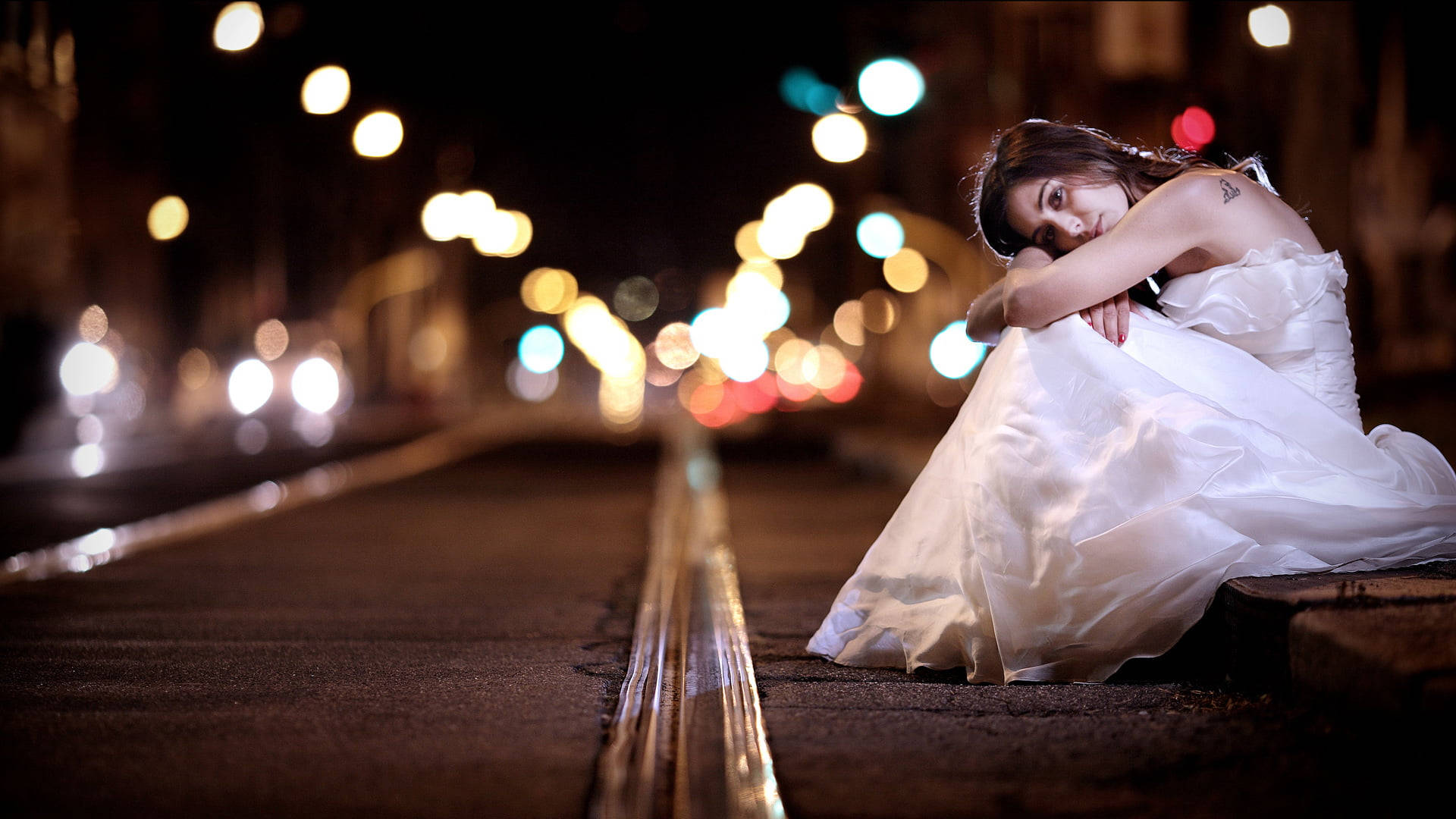 管狀婚紗 壁紙 — 一位身穿白色婚紗的女性夜晚獨自坐在城市街道上，被交通中閃爍的散景燈光包圍。