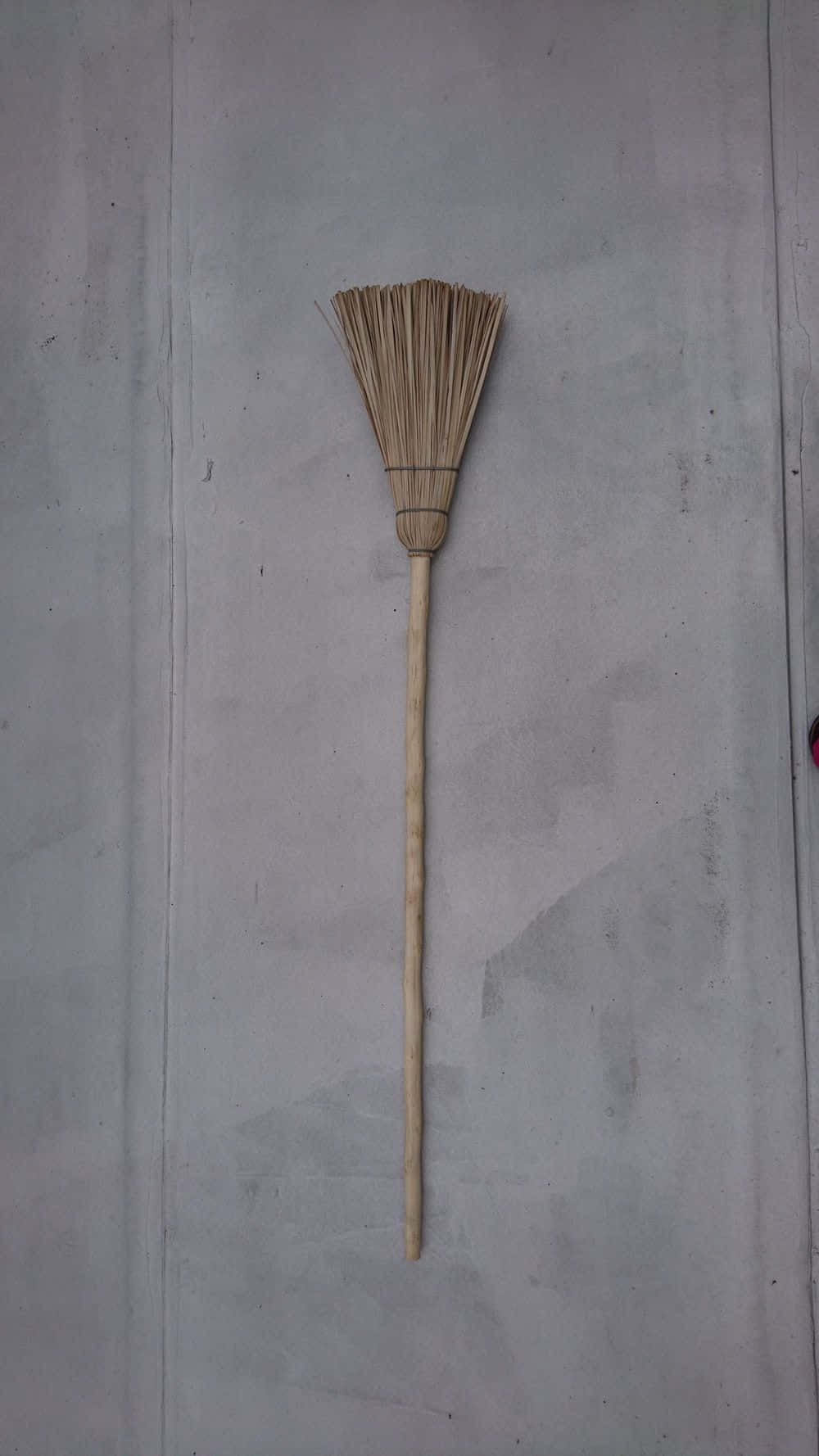 簡單掃帚圖 壁紙 — 一個簡單的稻草掃帚，帶有木柄，垂直掛在素色灰色混凝土牆上。