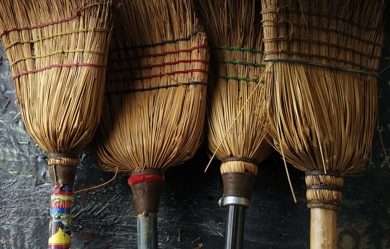 簡單而強大的家居工具：可靠的掃帚 壁紙 — 四把鄉村風格的掃帚，帶有編織稻草刷毛和彩色綁帶，掛在深色紋理牆上。