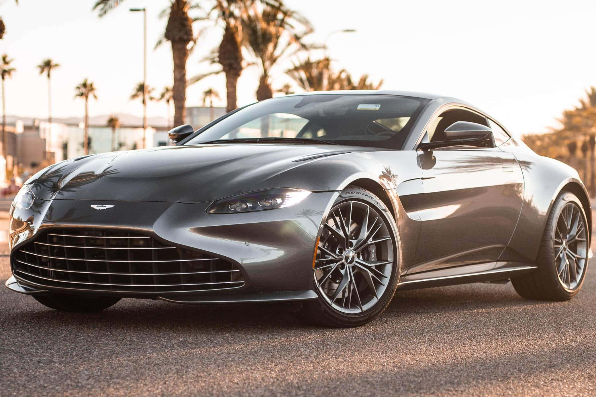 簡直令人驚嘆。 壁紙 — 一輛流線型的銀色Aston Martin跑車在金色夕陽下，在棕櫚樹旁的鋪砌道路上閃閃發光。