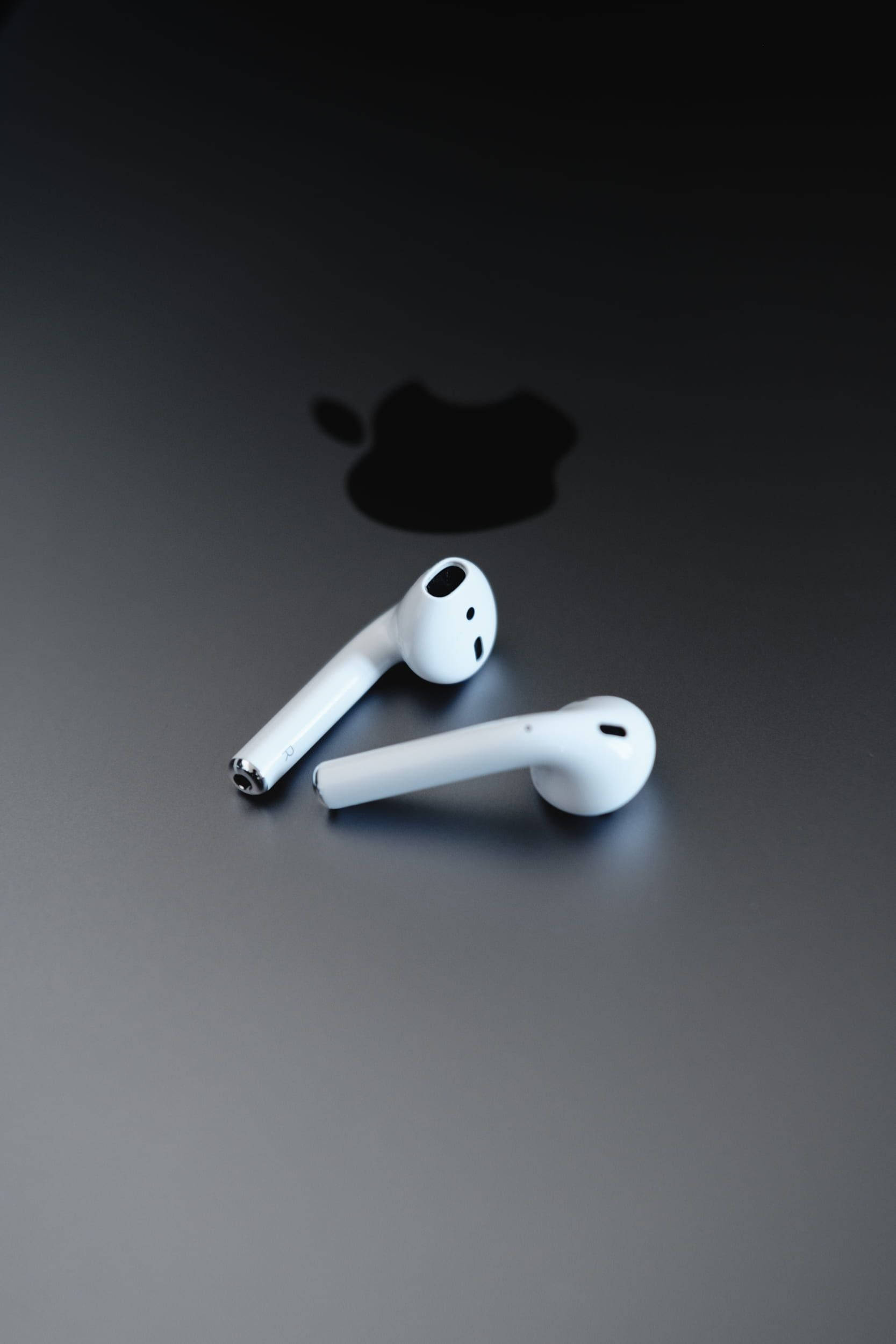 簡約 Apple AirPods 壁紙 — 兩個白色無線耳機放在深色表面上，背後有模糊的蘋果標誌陰影。