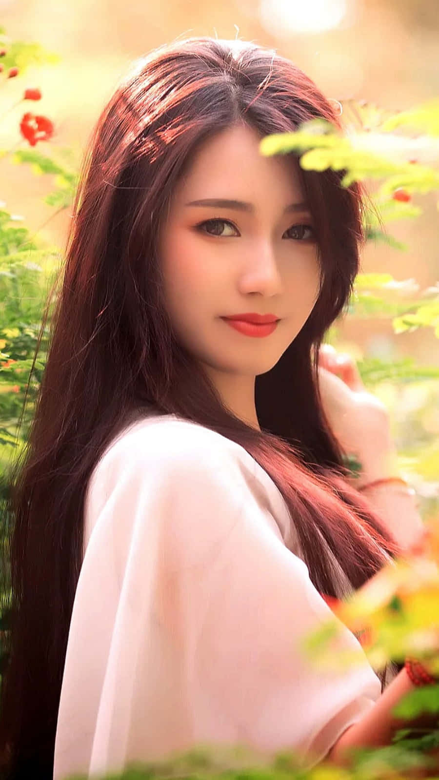 長黑髮、身穿白色上衣的女性站在陽光普照的綠植和紅花之間。