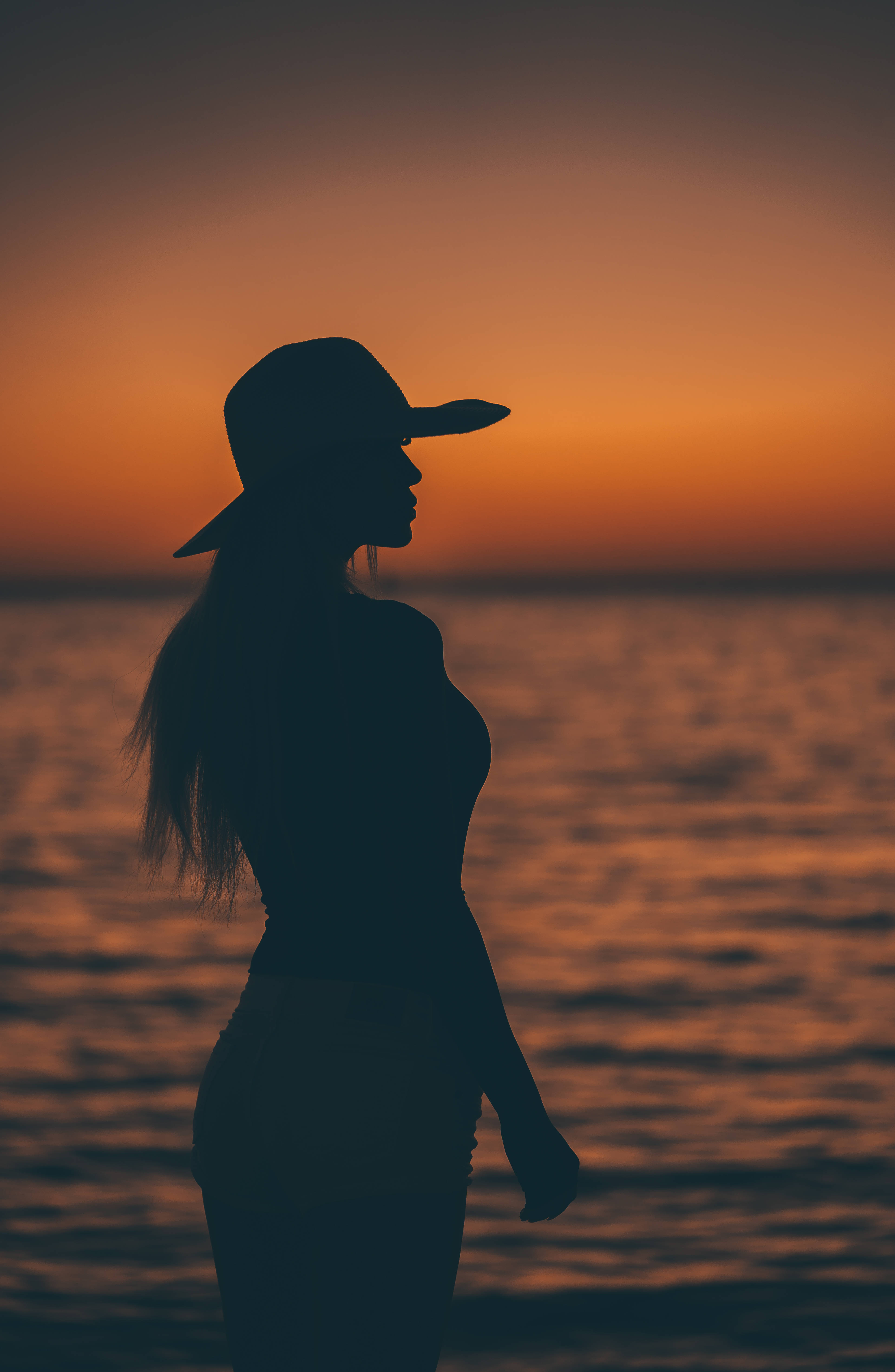 簡約暗黑風格戴帽女子夕陽 壁紙 — 一位戴寬邊帽的女子的剪影，在日落時分站在海邊，沐浴在溫暖的橙色光芒中。