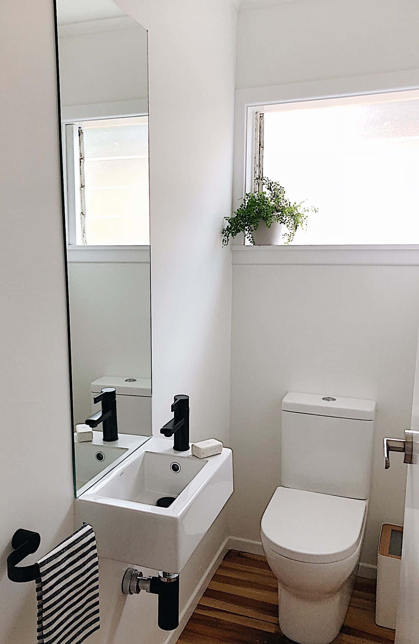 Toilet 桌布 壁紙 — 簡約浴室，配有白色潔具、黑色水龍頭、木地板和窗邊的綠色植物。