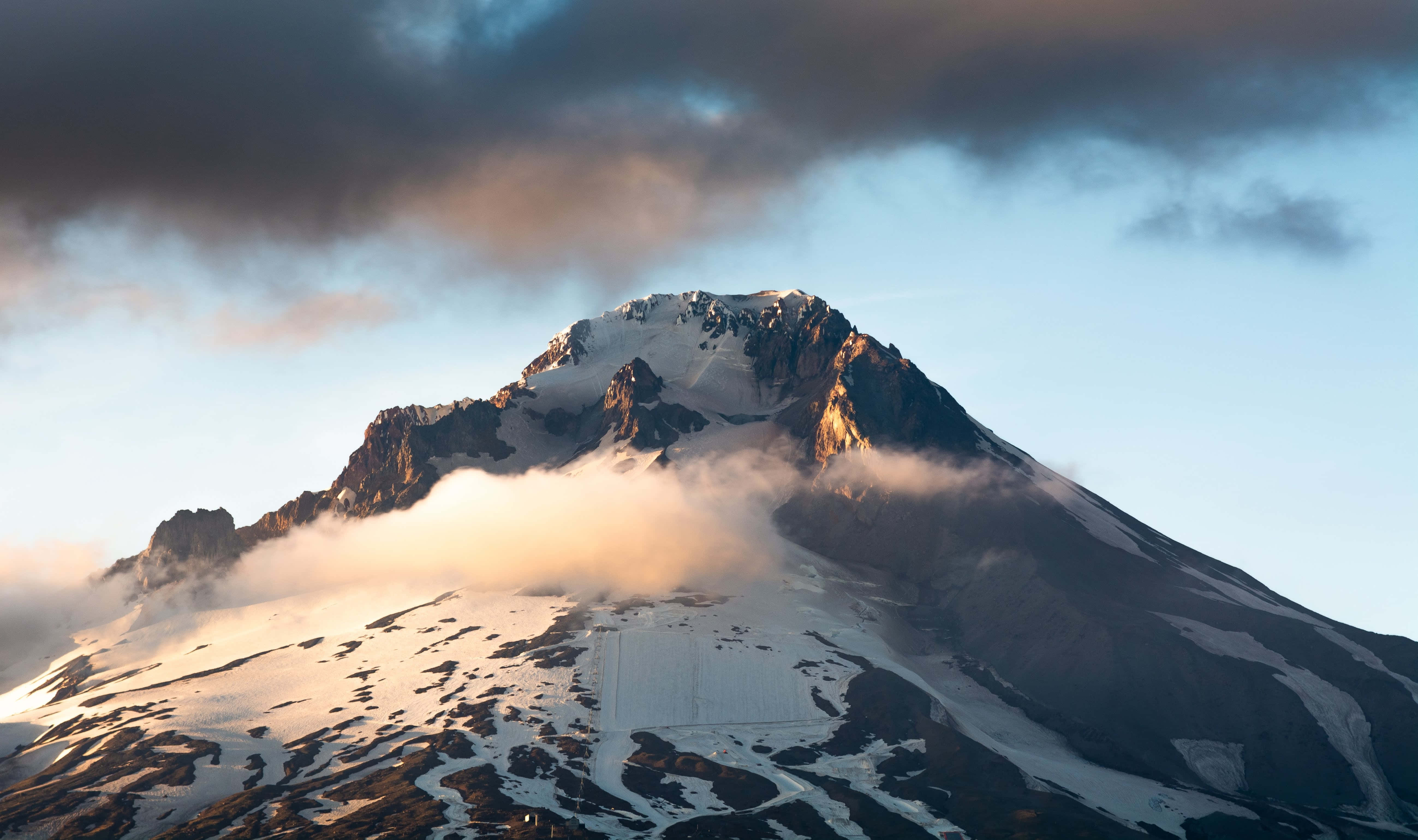 簡約藍天背景 壁紙 — 白雪覆蓋的山峰刺穿烏雲，金色的日出光芒照亮其崎嶇的山坡和飄浮的薄霧。