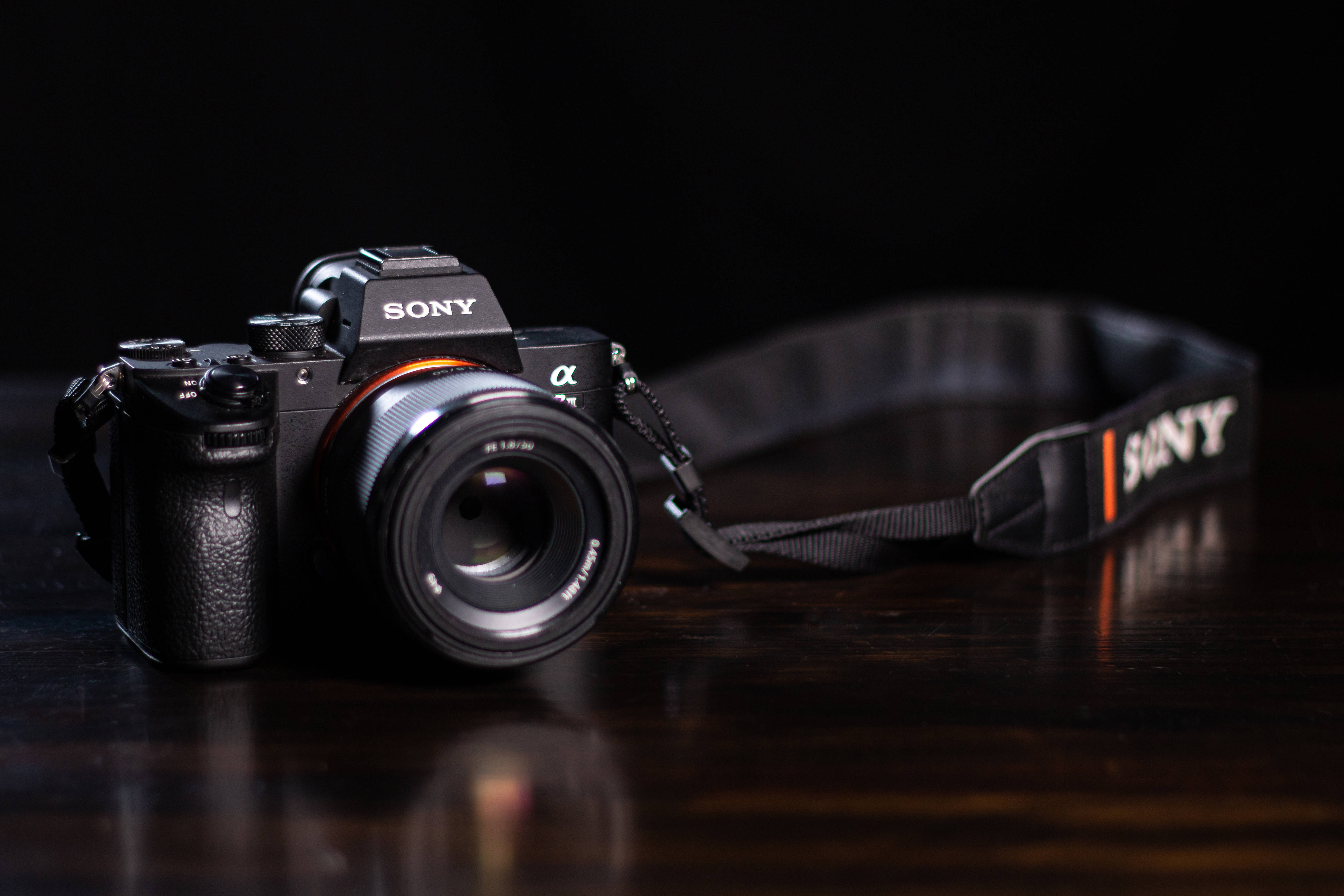 簡約高清索尼相機 壁紙 — 一部時尚的黑色Sony相機連鏡頭，放置在黑色背景前的深色木質表面上。
