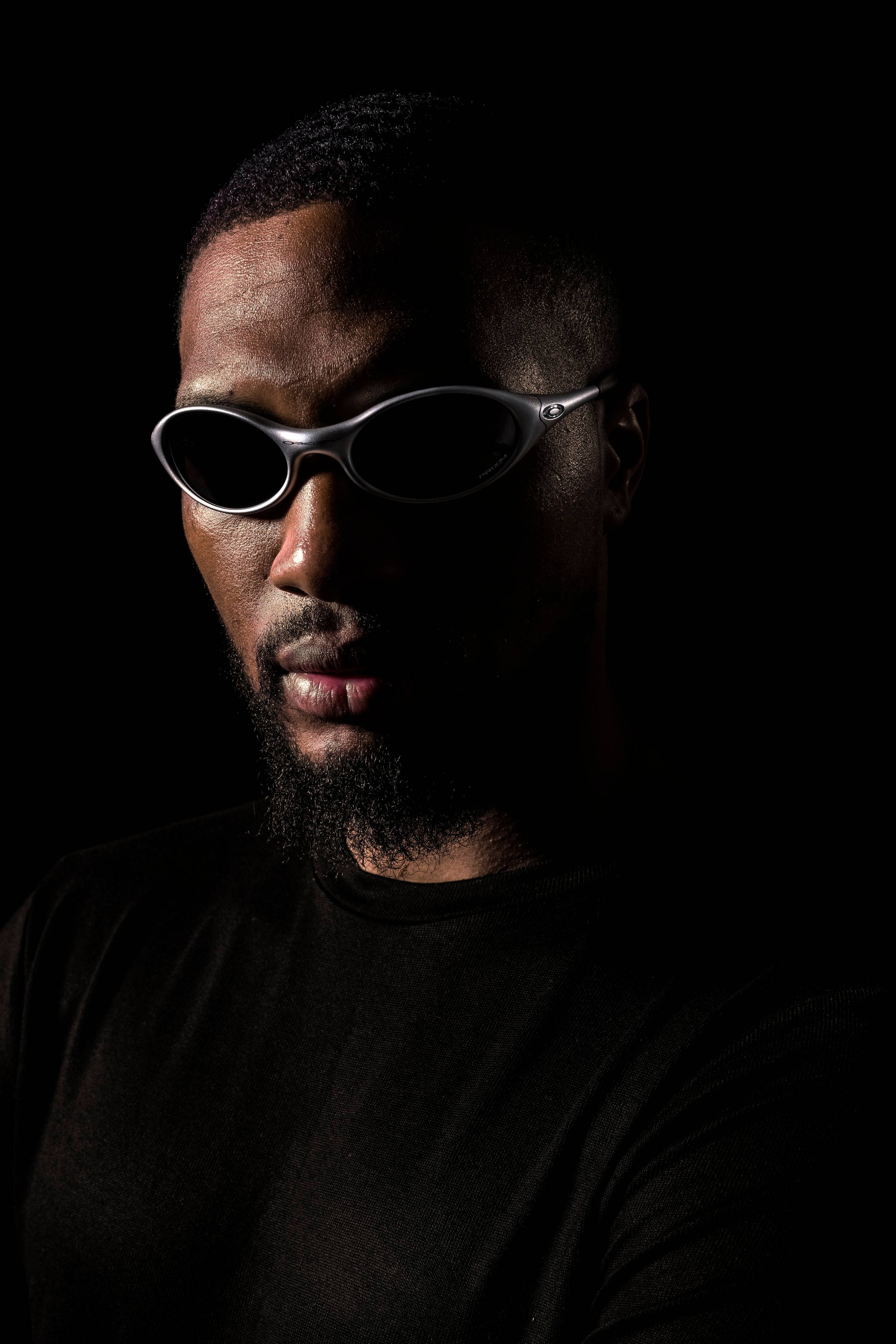 籃球員達米安·利拉德佩戴 Oakley 太陽眼鏡 壁紙 — 一個留鬍鬚嘅男人戴住時尚嘅銀色太陽眼鏡，背景係黑色，側面有強烈嘅燈光效果。