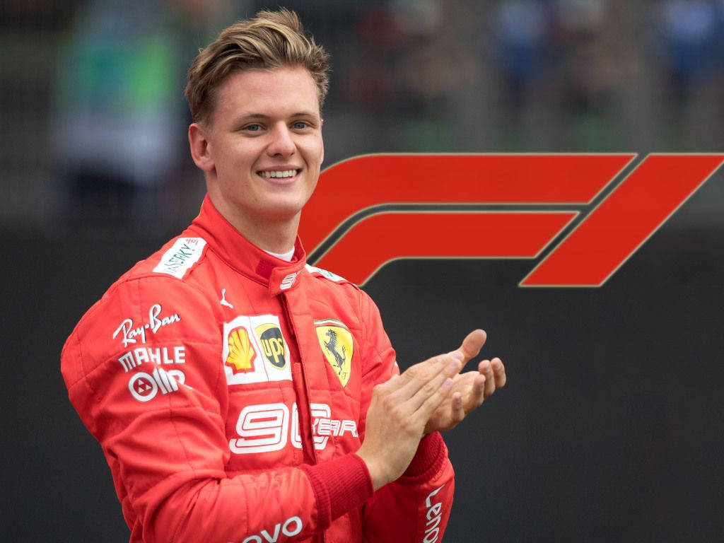 米克·舒馬赫自然肖像 壁紙 — 一位微笑的F1車手身穿紅色法拉利賽車服，在大型F1標誌前拍手。