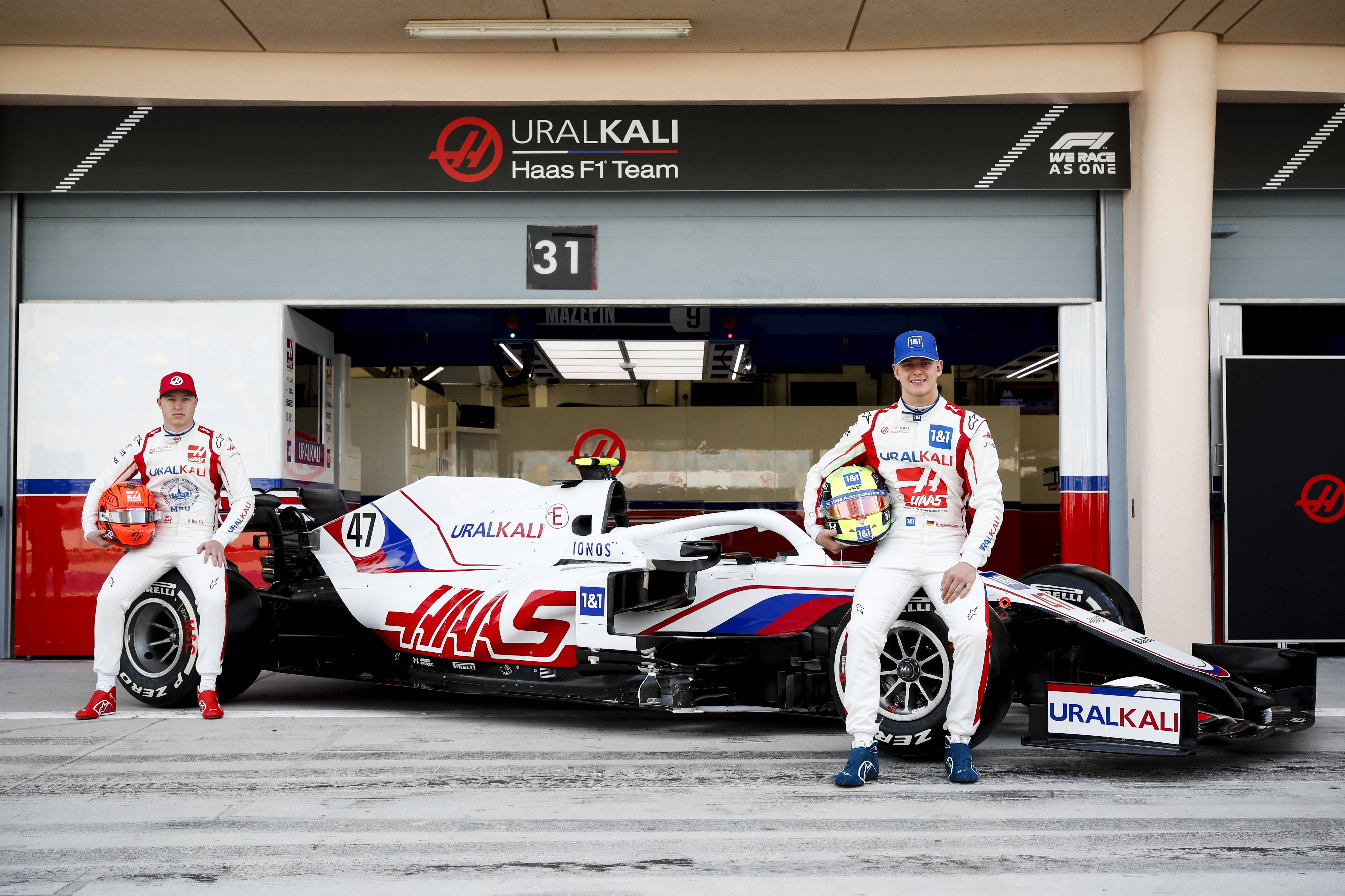 米克·舒馬赫與哈斯F1車隊 壁紙 — 兩名身穿白色賽車服的F1車手在車庫中自信地站在他們的Haas F1車隊賽車旁。