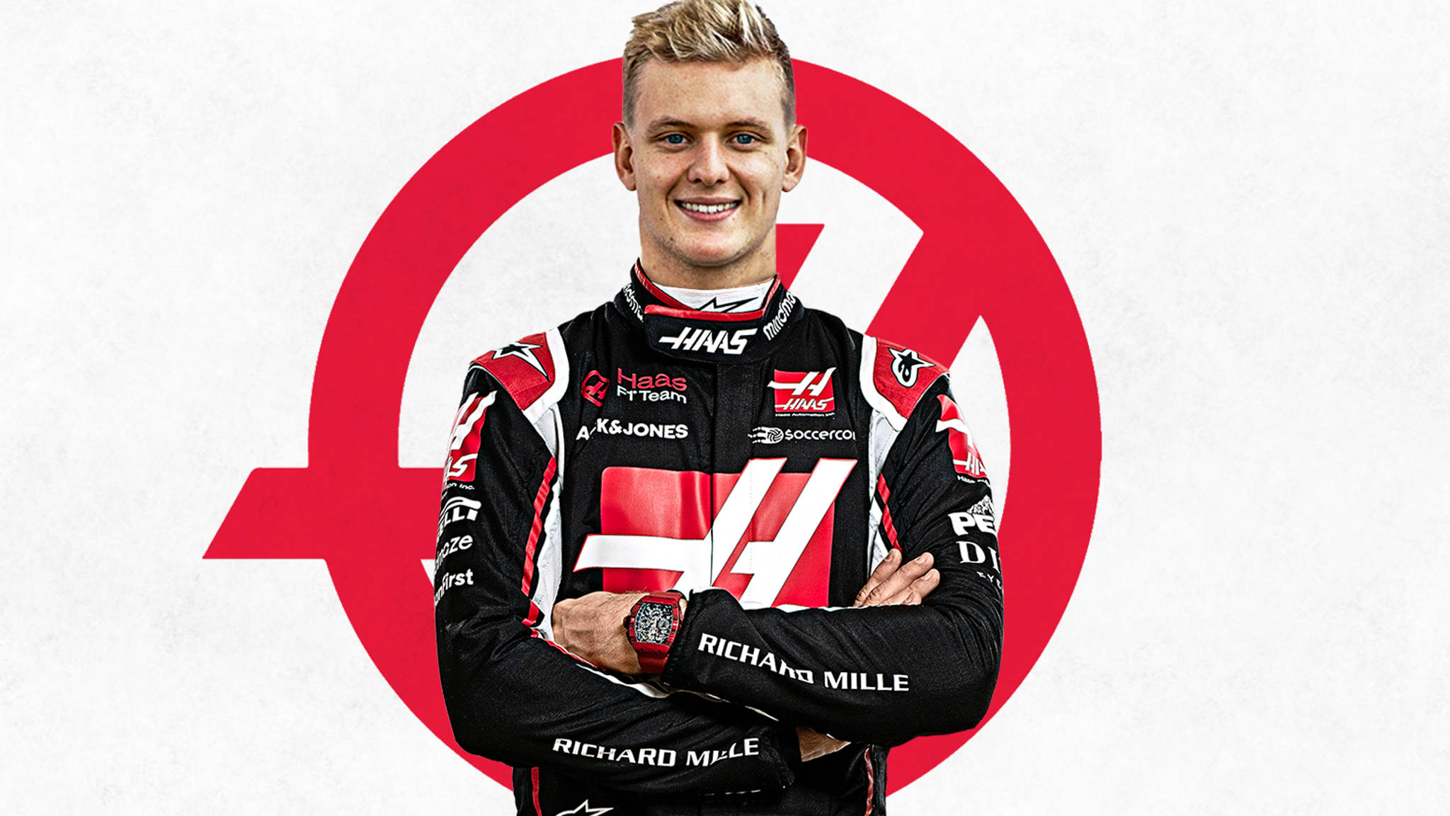 米克·舒馬赫雙臂交叉肖像 壁紙 — 一位面帶微笑的F1車手身穿黑紅色的Haas賽車服，雙臂交叉，自信地站在白色背景上一個醒目的紅色圓形標誌前。