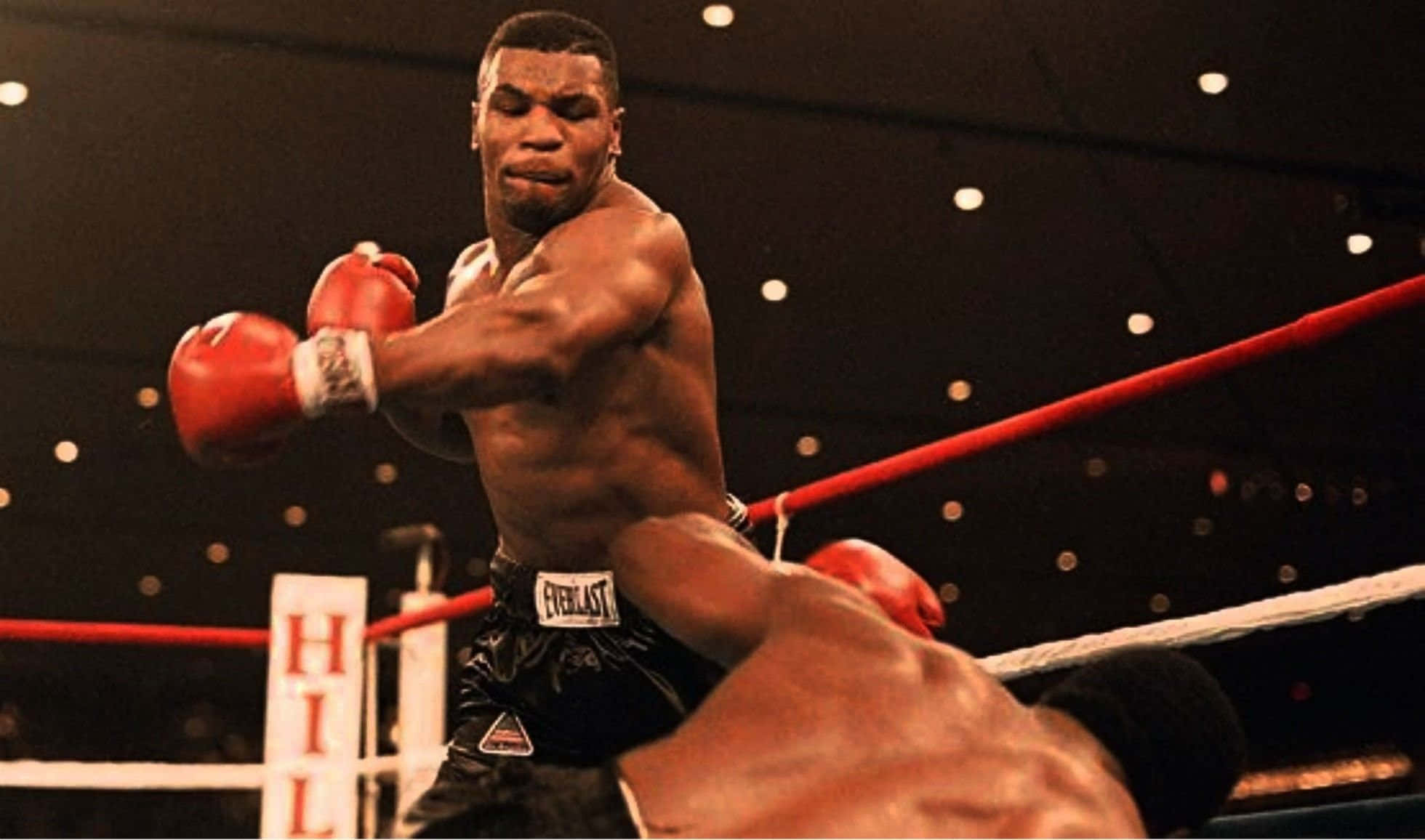 mike tyson 桌布 壁紙 — 米克泰臣在昏暗的拳擊擂台上，背景有紅色繩索和模糊的人群燈光，打出強力一拳。