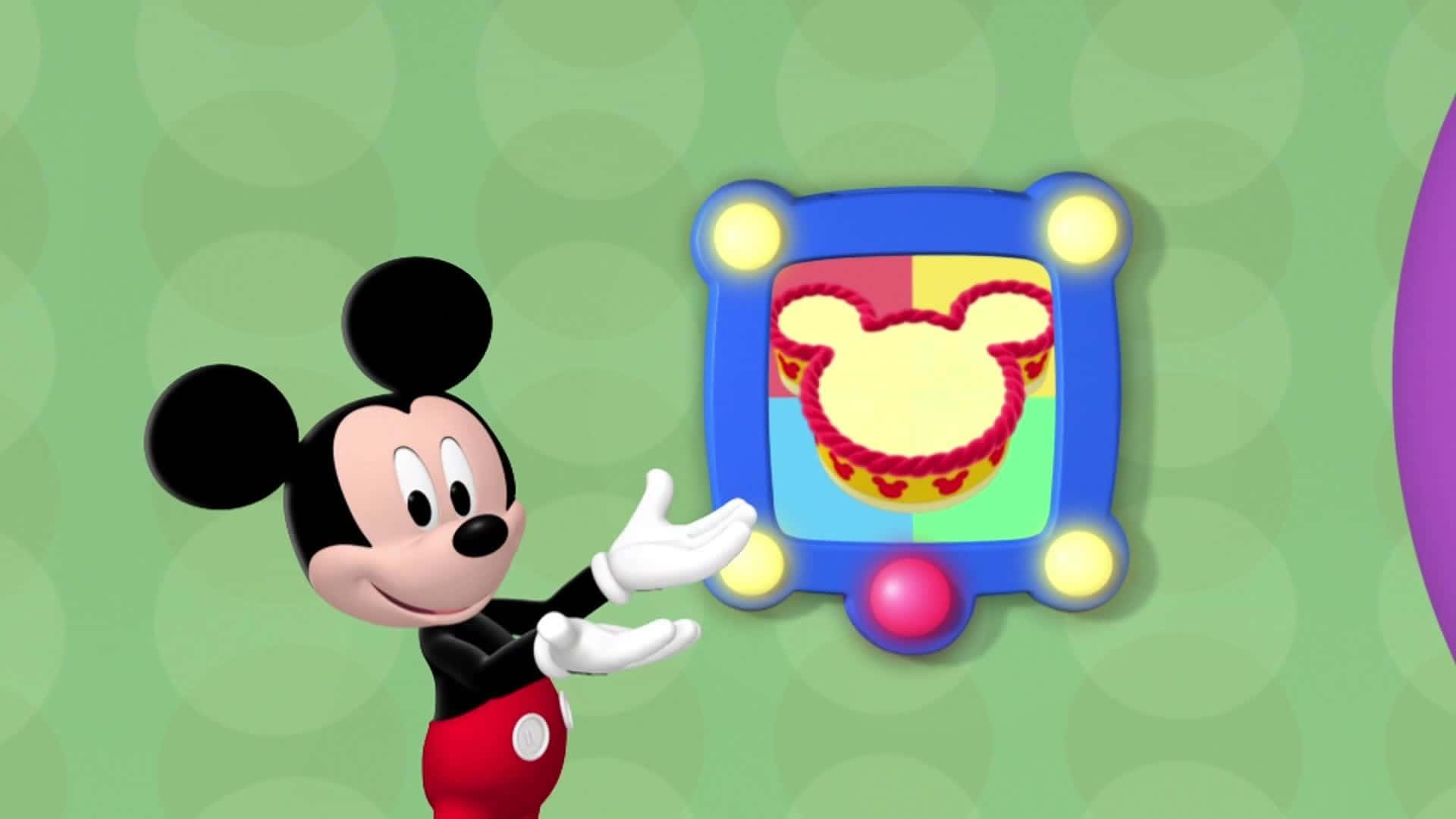 Mickey Mouse Clubhouse 桌布 壁紙 — 米奇老鼠穿著經典紅色短褲，指向一個色彩繽紛、鑲框的米奇頭形蛋糕，上面有紅繩和心形裝飾。