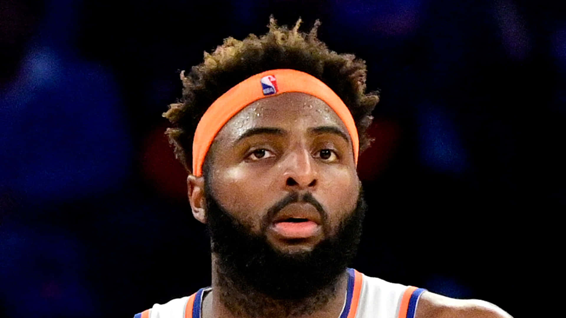 米契爾·羅賓遜NBA球員照 壁紙 — 專注的籃球運動員特寫，留鬍鬚，戴橙色頭帶，在明亮的場館燈光下比賽。