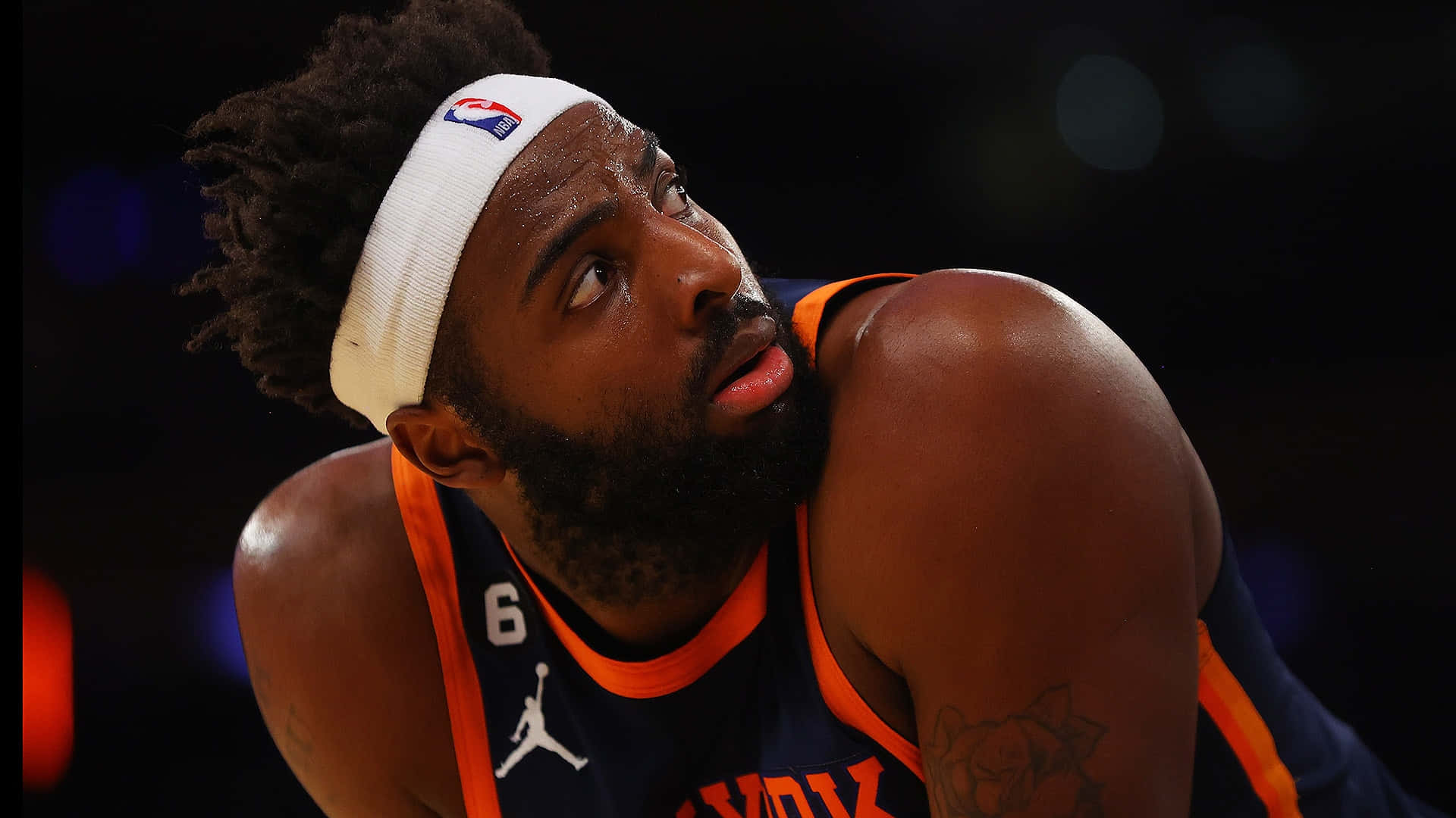 米契爾·羅賓遜NBA籃球球員 壁紙 — 一位身穿鳳凰城太陽隊球衣的專注籃球運動員，在場館燈光下身體前傾，目光銳利。