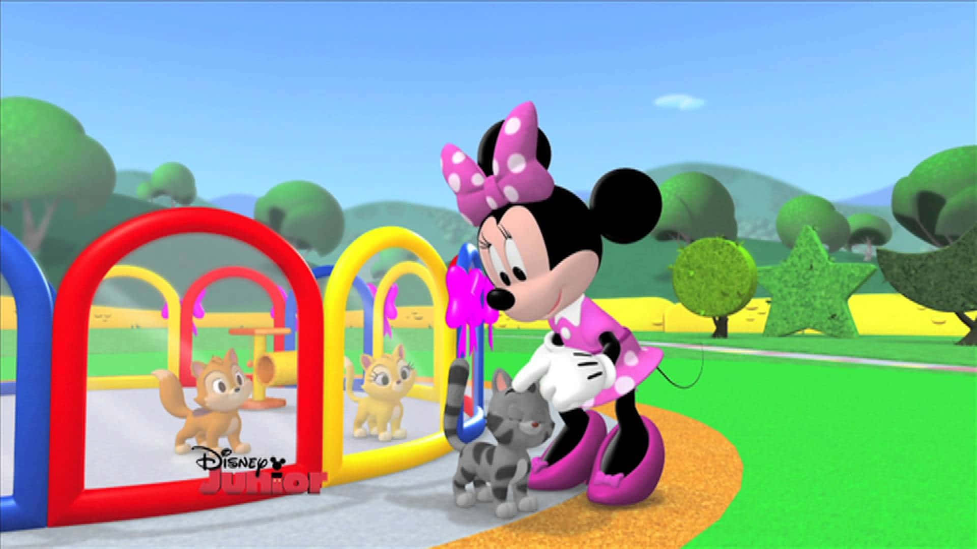 Mickey Mouse Clubhouse 桌布 壁紙 — 米妮老鼠身穿粉紅色圓點裙和蝴蝶結，在明亮的藍天下，在色彩繽紛的遊樂場拱門附近與小貓玩耍。