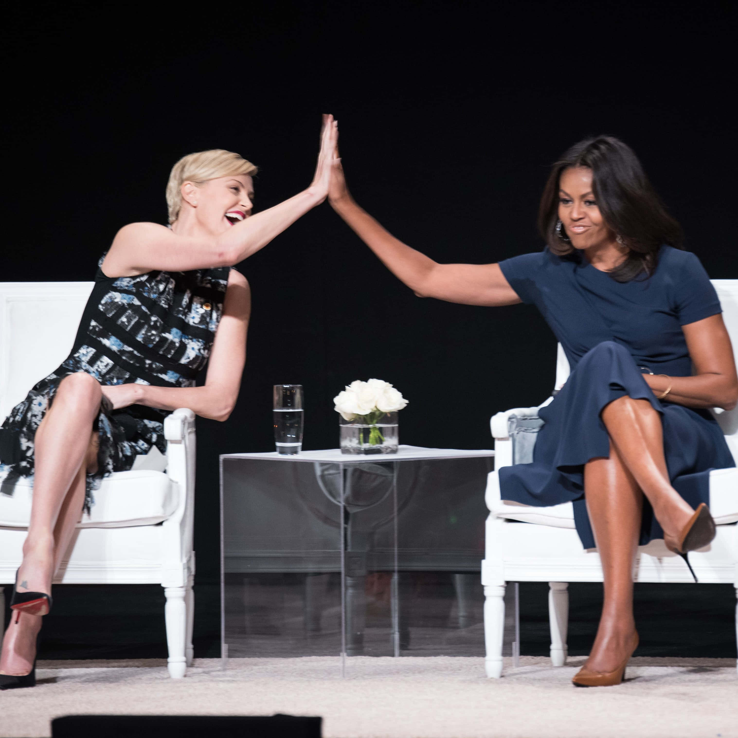 米歇爾·奧巴馬與查理茲·塞隆高五圖片 壁紙 — 兩位女士坐在黑暗舞台上的白色椅子上，中間有一張玻璃桌，愉快地擊掌。