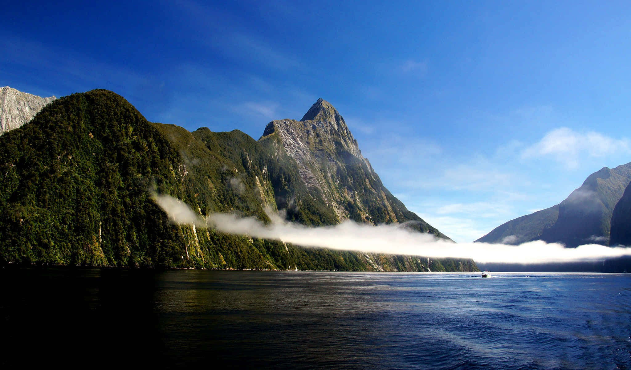 米爾福德峽灣雲線美景 壁紙 — 寧靜的峽灣，霧氣繚繞著高聳的綠色山脈，清澈的藍天下，平靜的水面上有一艘小船。