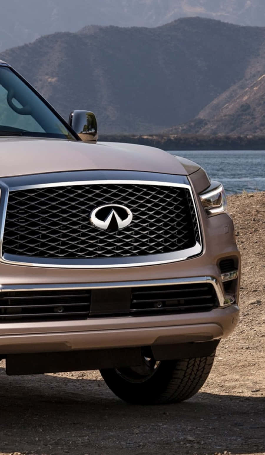 Infiniti Qx80 桌布 壁紙 — 米色Infiniti SUV前格柵的特寫，停在水邊，背景有山。