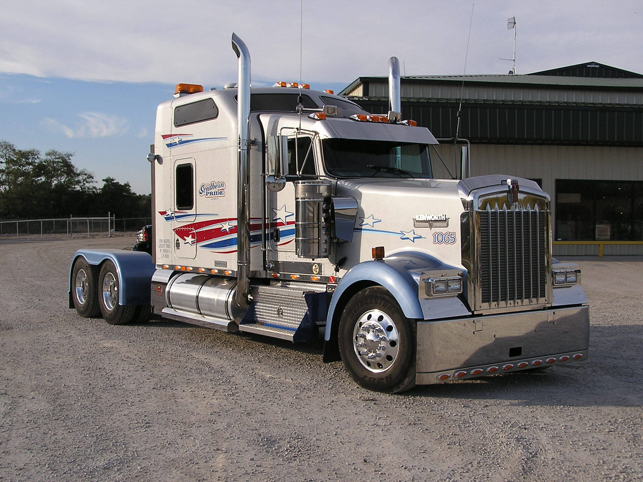 米色Kenworth W900 壁紙 — 一輛閃亮的銀色 Kenworth 半掛卡車，帶有愛國紅白藍貼花，停在倉庫附近的礫石上。