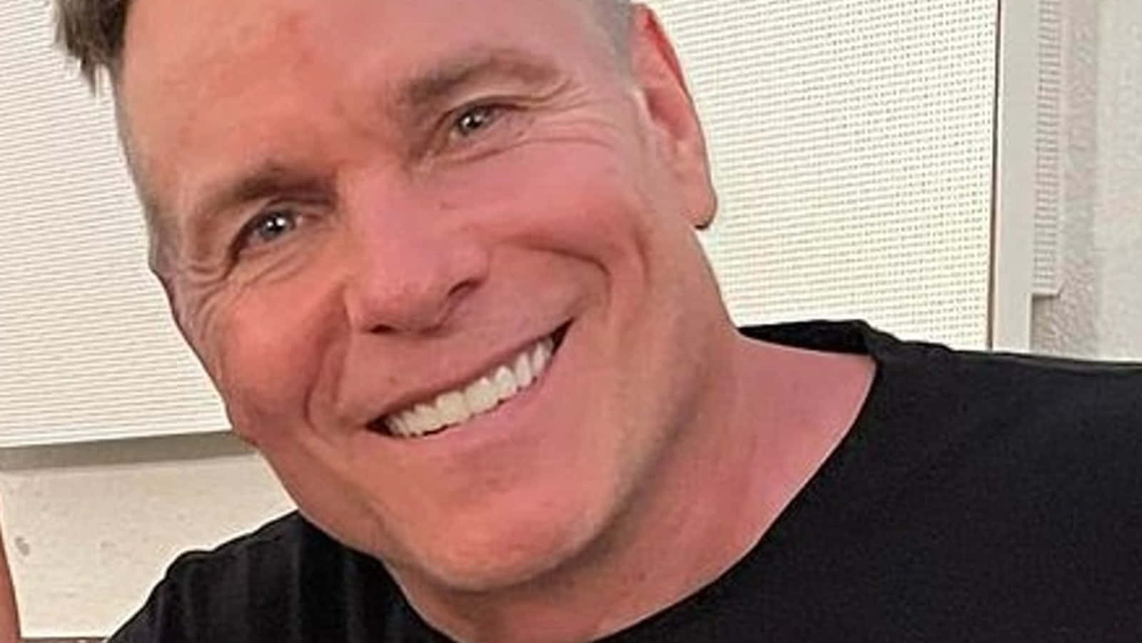 米色背景下的蘭尼·波福 壁紙 — 一張特寫照片，一位短髮微笑的男子穿著黑色襯衫，背景是白色紋理牆壁。