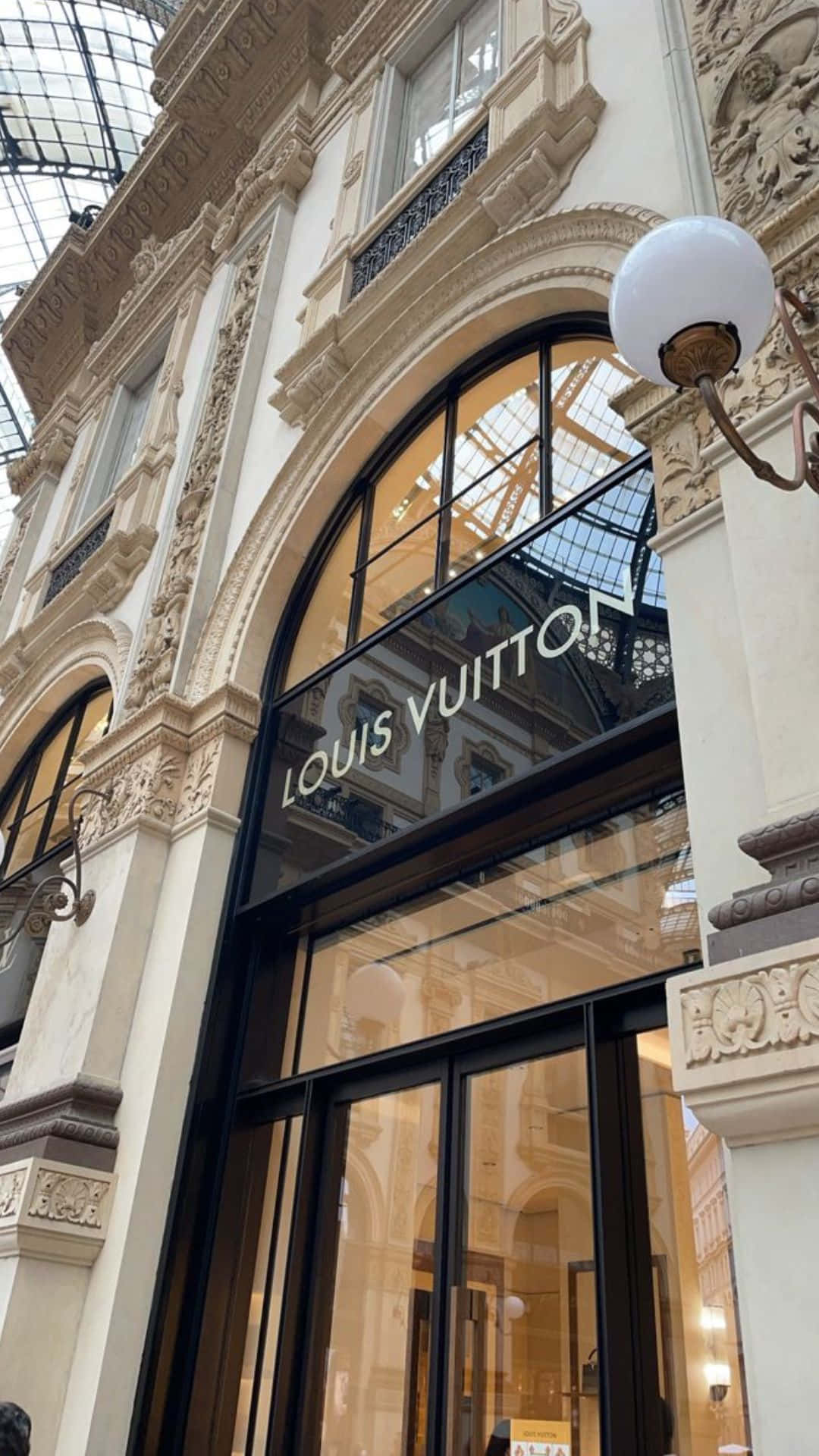 米色香草路易威登門面 壁紙 — 宏偉的Louis Vuitton精品店入口，上方是華麗的玻璃天花，飾有精緻的石雕和溫暖的室內燈光。