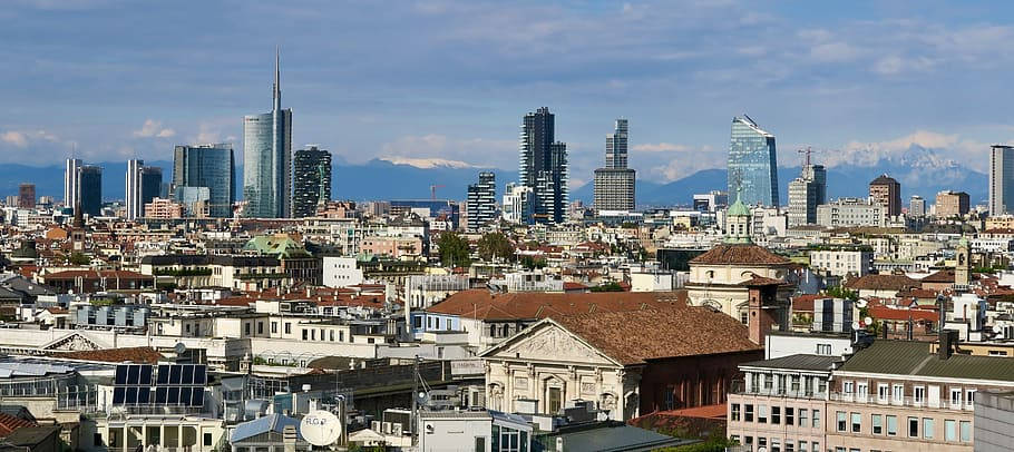 米蘭城市景觀，現代塔樓和歷史建築在藍天下，遠處有山脈。