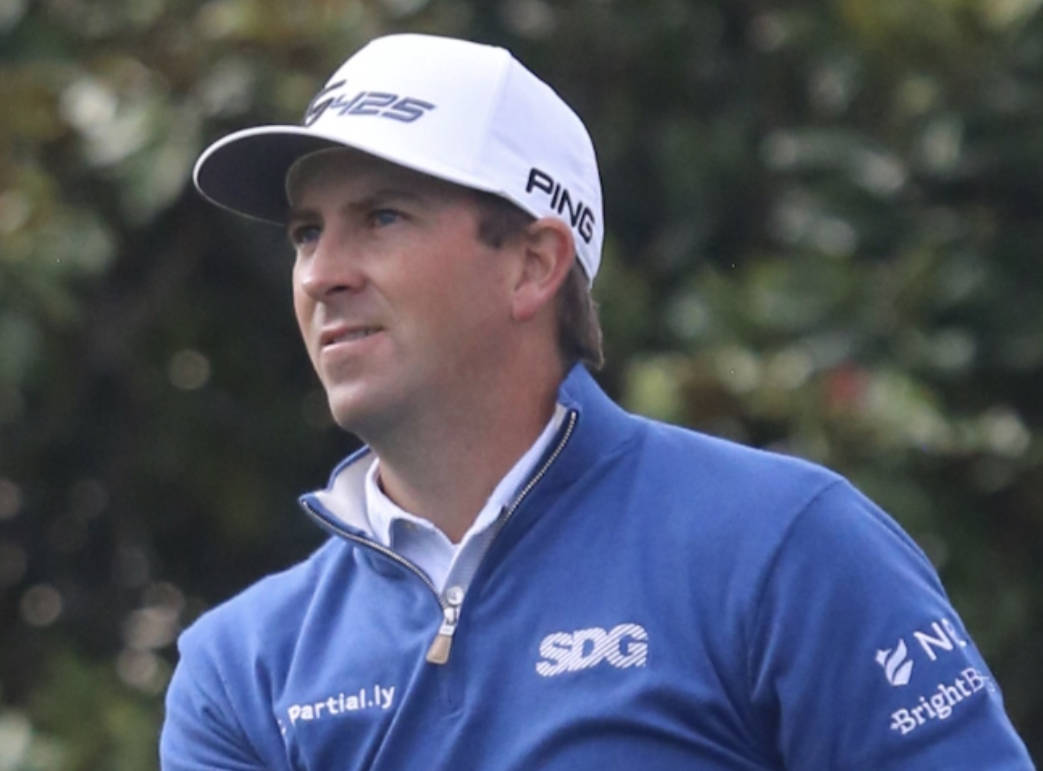 米高·湯普森身穿藍色上衣 壁紙 — 一位專注的高爾夫球手，身穿藍色外套和白色帽子，站在模糊的綠色樹葉背景前。
