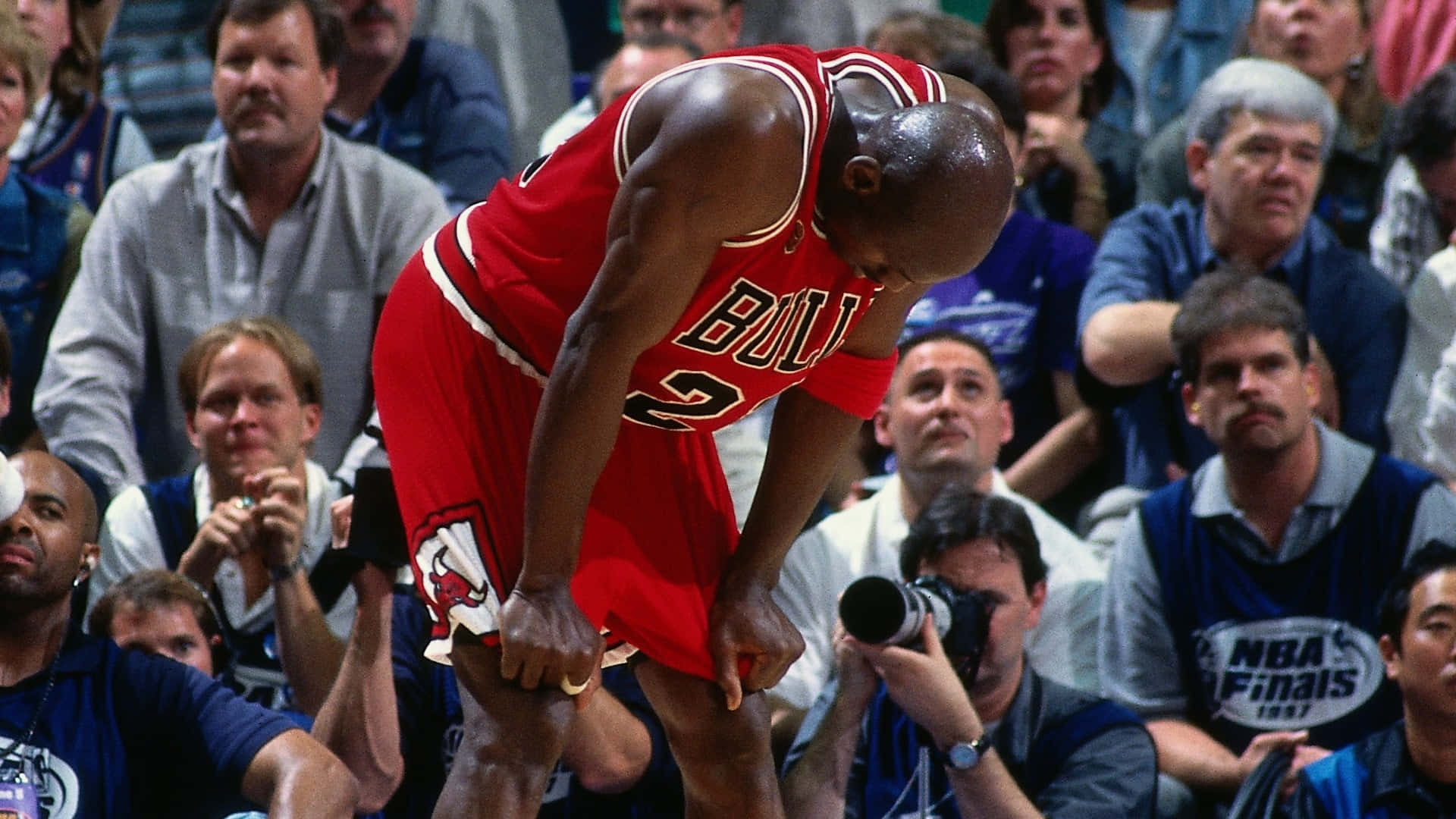 Jordan 桌布 壁紙 — 米高佐敦身穿紅色芝加哥公牛隊球衣，在NBA總決賽比賽中彎腰向前，筋疲力盡，背景有觀眾。