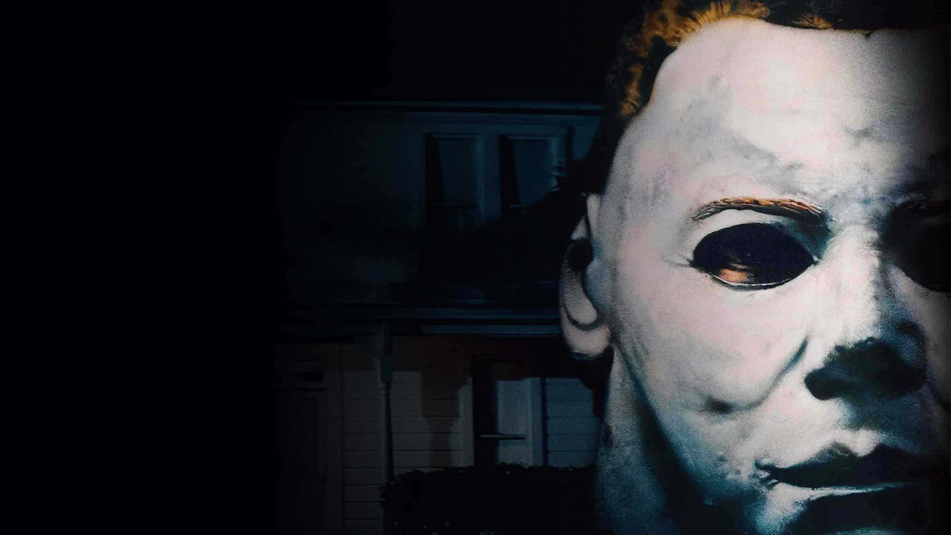 Mike Myers 桌布 壁紙 — 米高邁亞斯標誌性白色面具的特寫，眼睛深邃，背景是夜間燈光昏暗的郊區房屋。