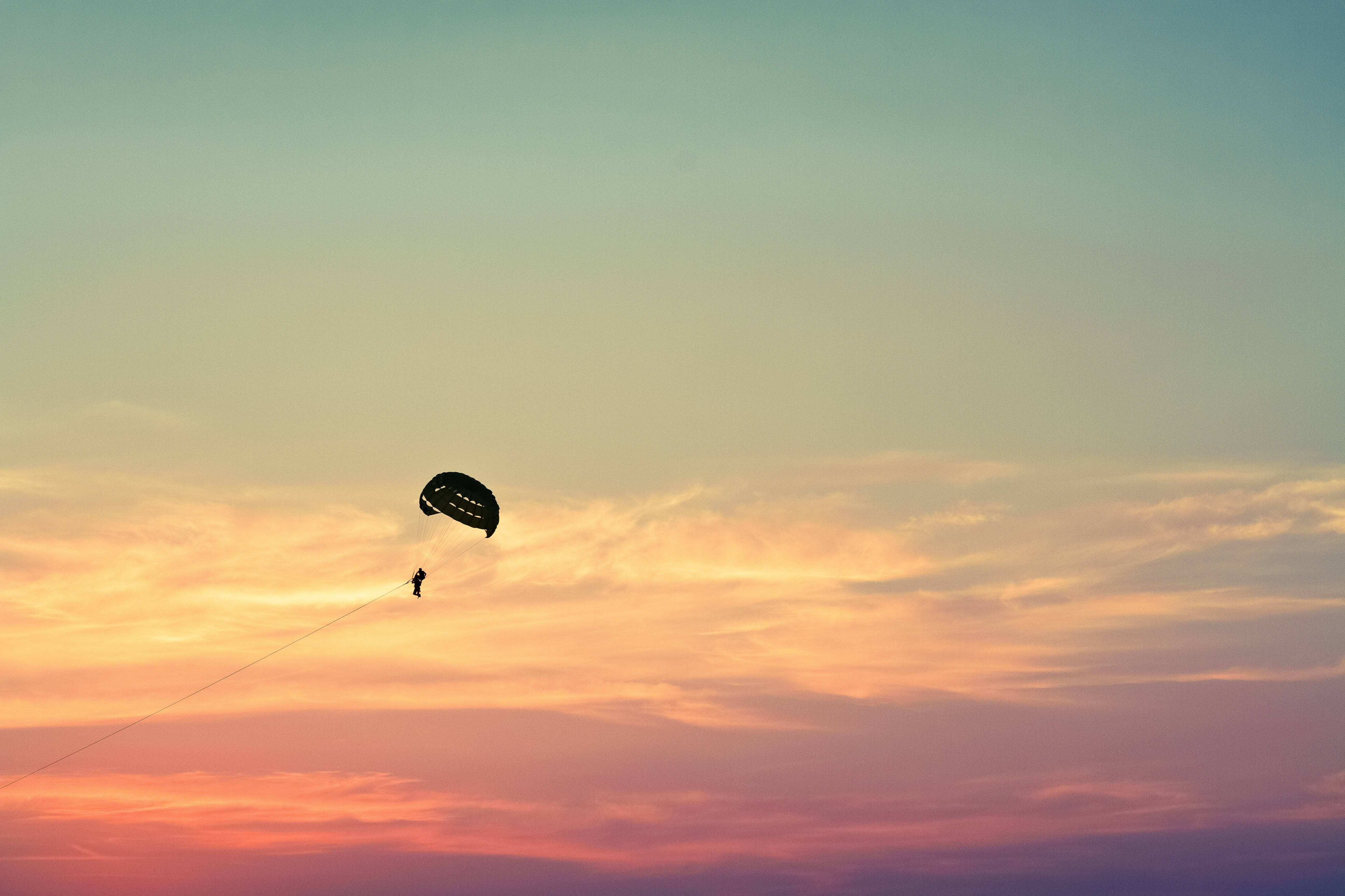 粉彩夕陽下傘降飛行 壁紙 — 滑翔傘剪影映襯著充滿活力的日落天空，帶有橙色、粉紅色和藍綠色的雲彩條紋。