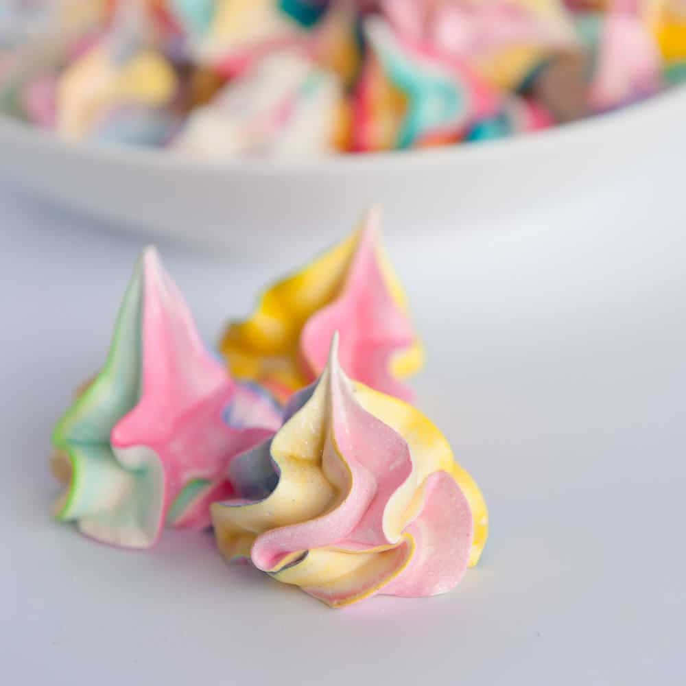 Meringue 桌布 壁紙 — 粉紅、黃色和藍綠色的柔和馬卡龍曲奇在白色表面上呈現鮮豔漩渦，背景有一個模糊的碗。
