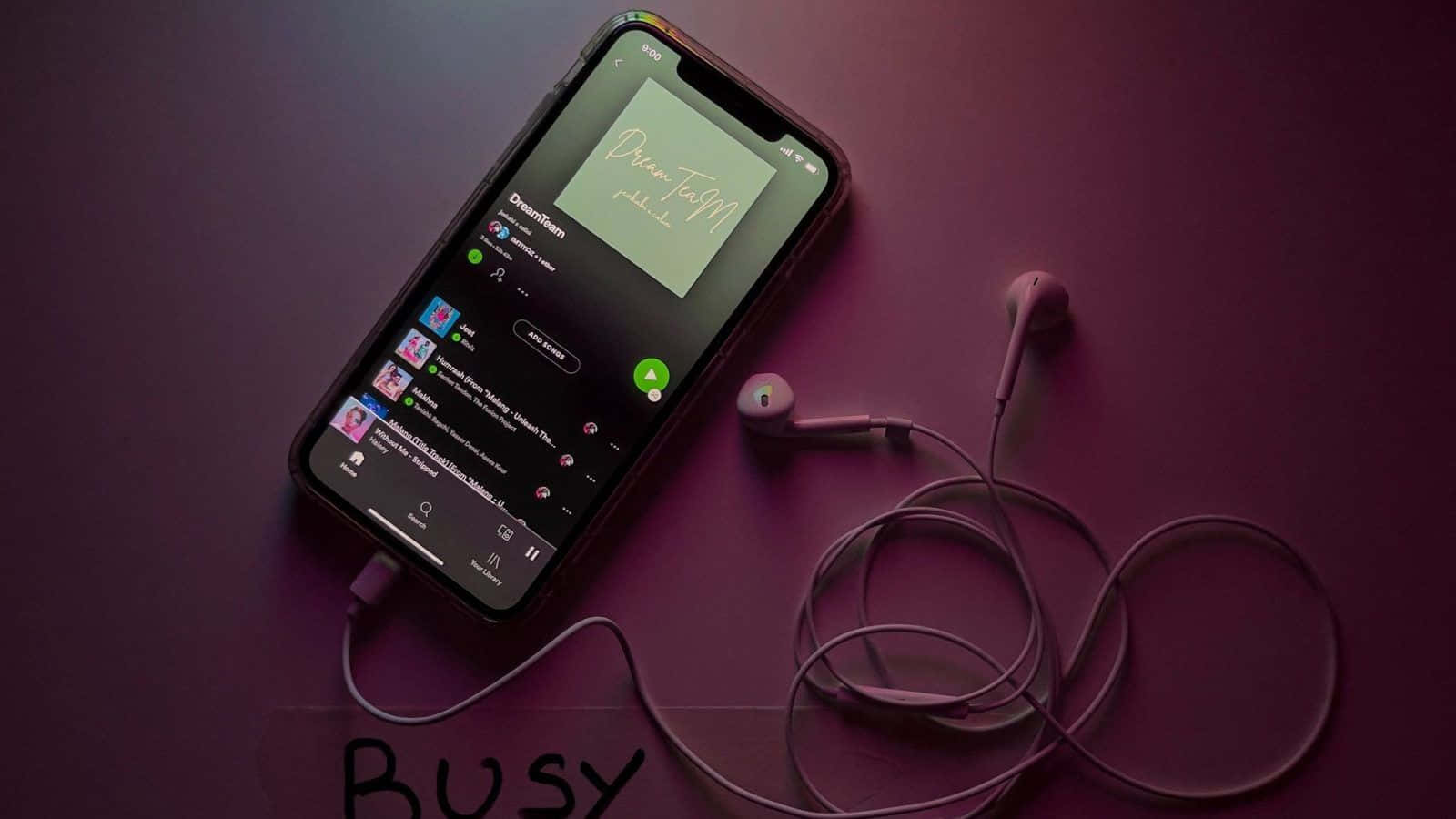 智能手機帶音樂應用程式和耳機，放在紫色表面上，下方寫有「BUSY」。 — 粉紅色 iPhone 壁紙