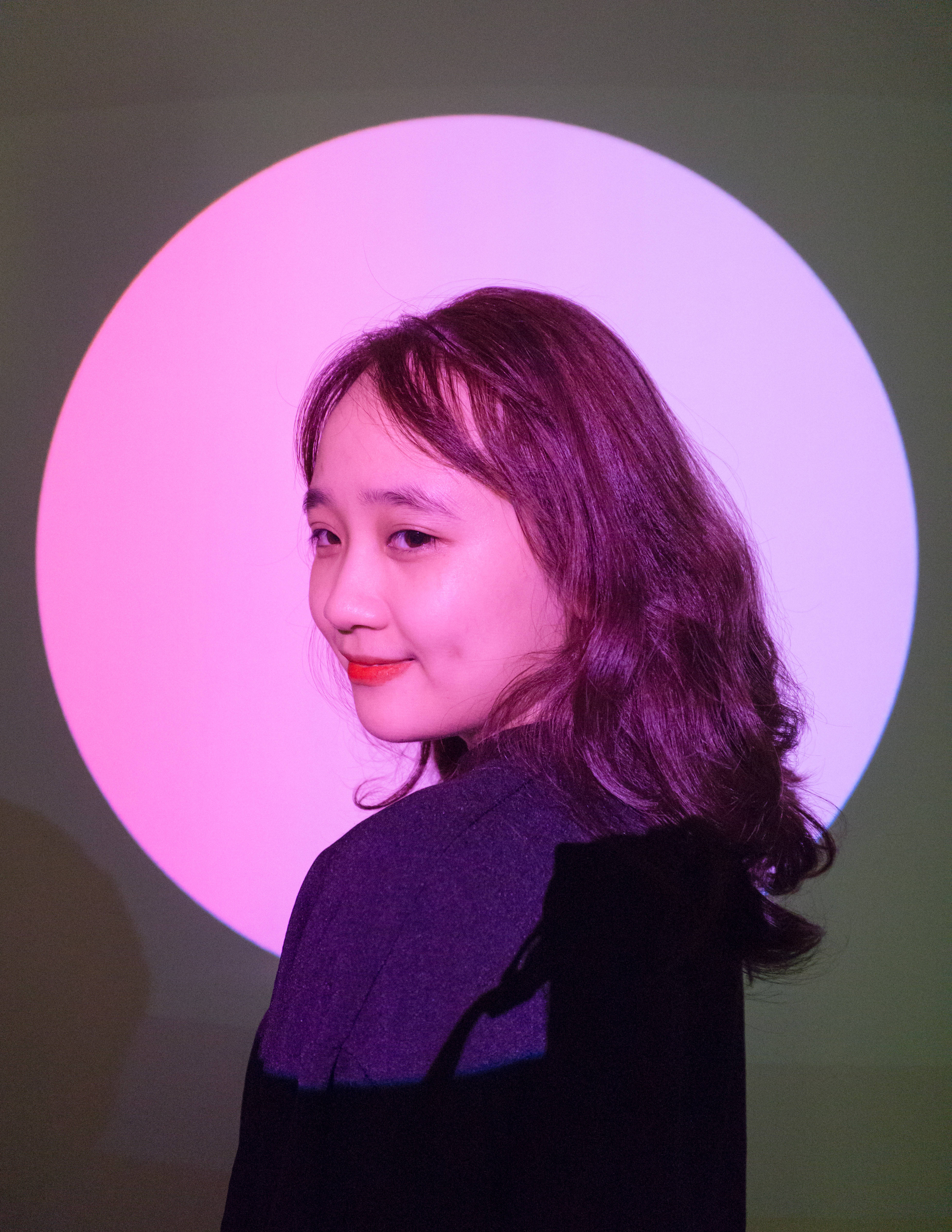 粉紅光圈中的可愛女孩 壁紙 — 一位年輕女性留著波浪長髮，在發光的粉紅色圓形背景前溫柔微笑。