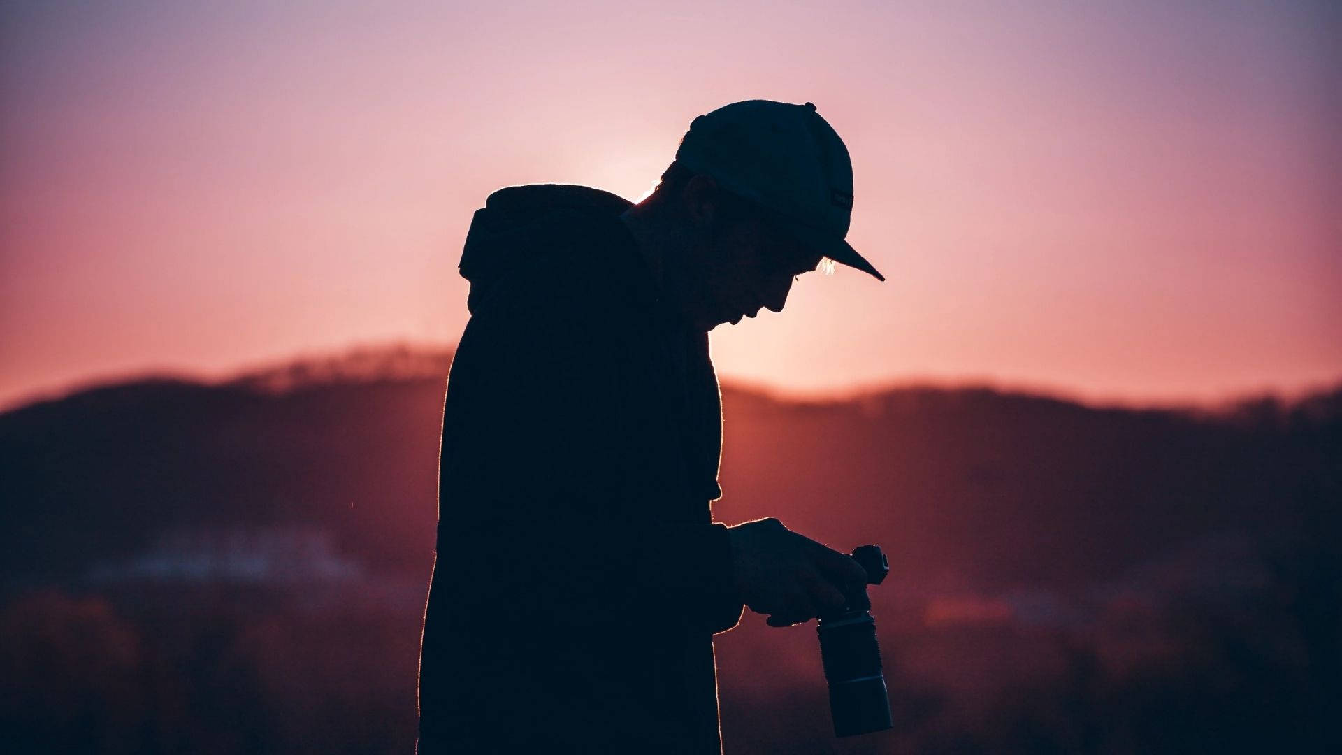 粉紅日落下的攝影師 壁紙 — 一個人戴著帽子和連帽衫的剪影，在遠山上方鮮豔的粉紅色和橙色日落背景下調整相機。