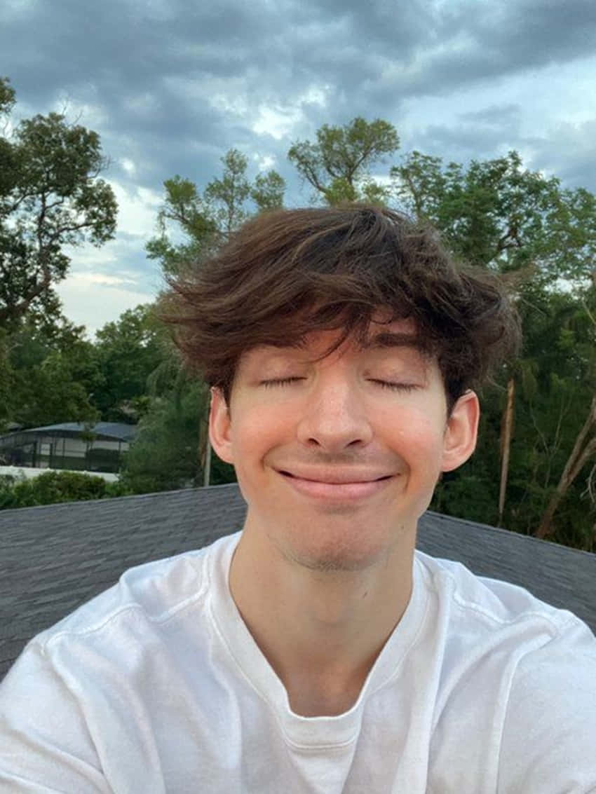 年輕男子在戶外陰天下閉眼微笑，背景有樹木。