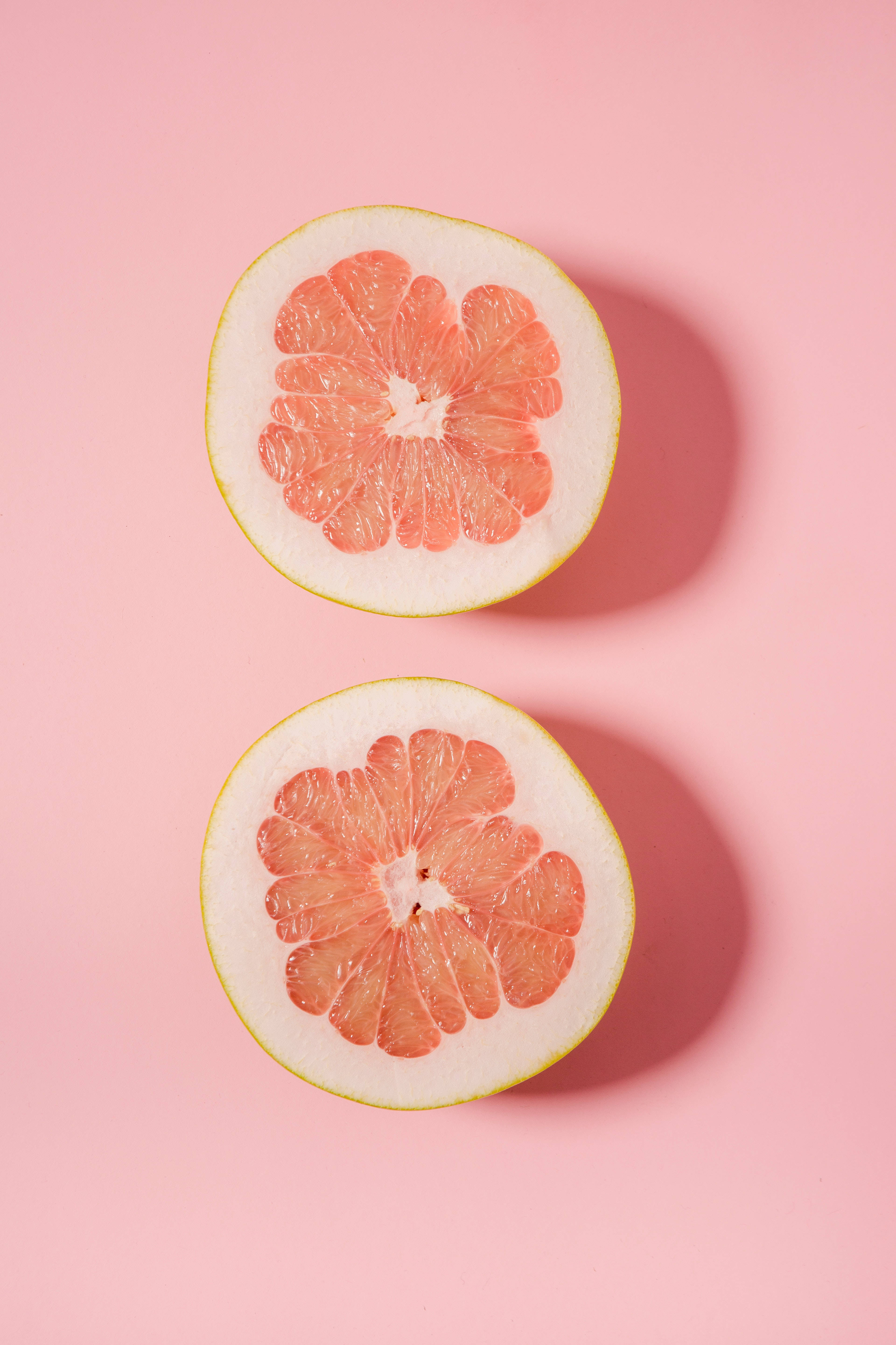 粉紅背景上的 pomelo 壁紙 — 兩個粉紅西柚切開一半，露出多汁的果肉，襯托柔和的粉紅色背景。