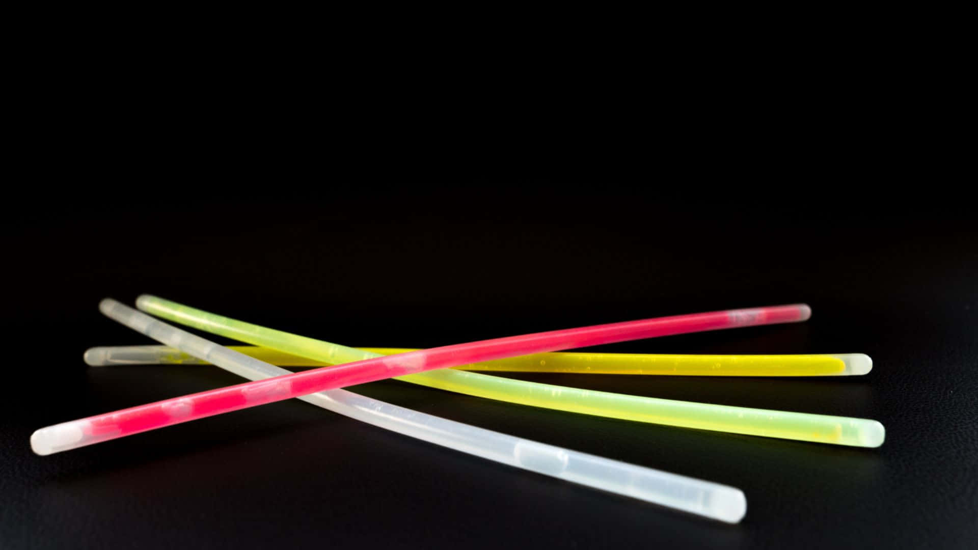 Glow In The Dark 桌布 壁紙 — 粉紅色、黃色和白色的鮮豔發光棒在純黑色背景上交錯。