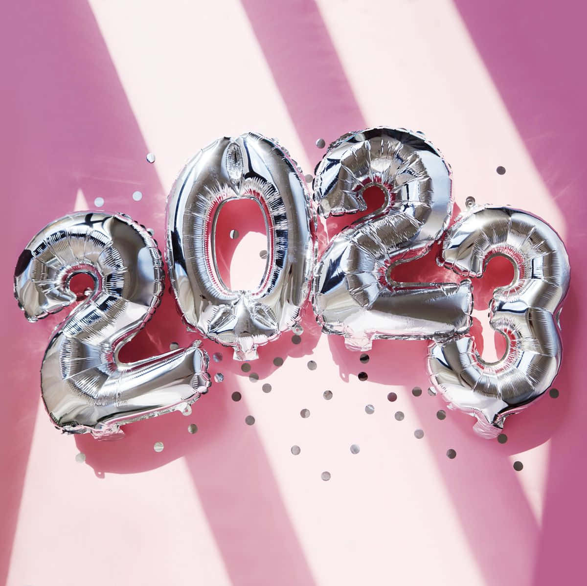 粉紅色2023新年氣球圖 壁紙 — 閃亮銀色2023數字氣球放置在粉紅色表面上，周圍散落著五彩紙屑和戲劇性的光線。