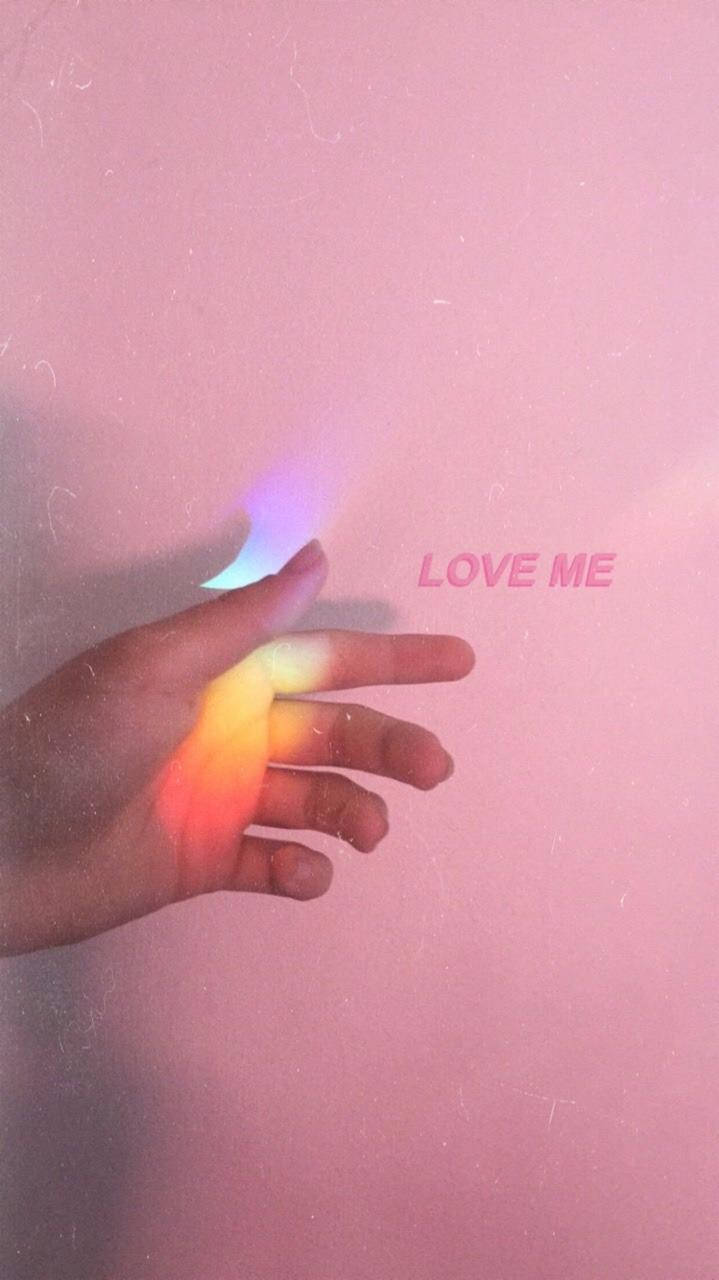 手在粉紅色牆上投射彩虹光，帶有「LOVE ME」文字。