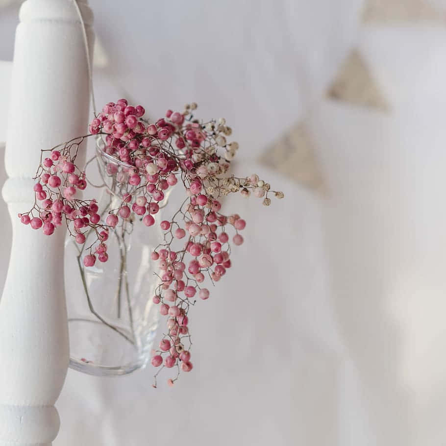 Pink Peppercorn 桌布 壁紙 — 粉紅色乾燥莓果從一個透明玻璃花瓶傾瀉而出，花瓶放在白色木製紡錘上，背景是柔和的白色牆壁，帶有模糊的三角形裝飾。