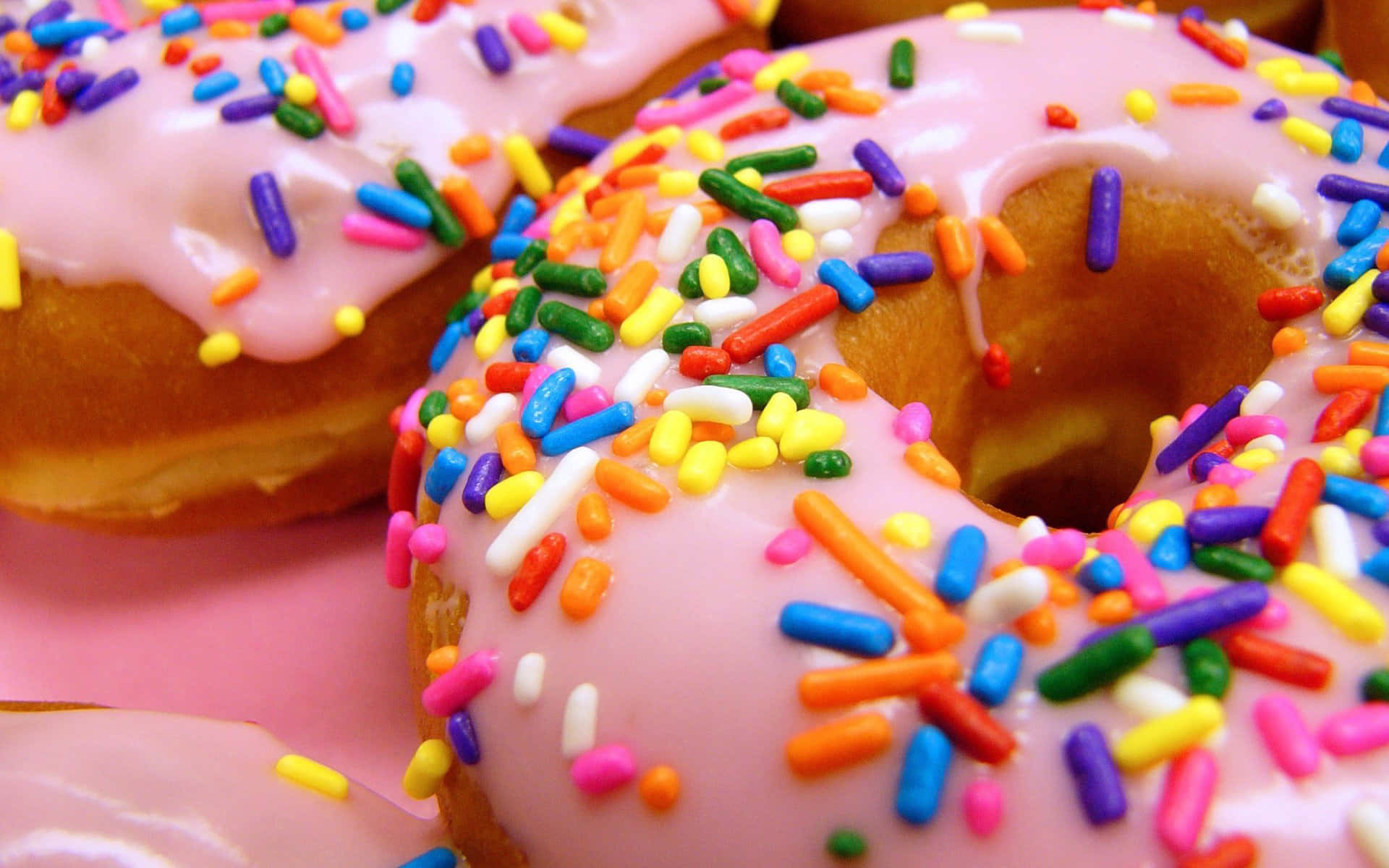 Cute Donut 桌布 壁紙 — 粉紅色表面上覆蓋著彩色彩虹糖霜的光澤粉紅色糖霜甜甜圈特寫。