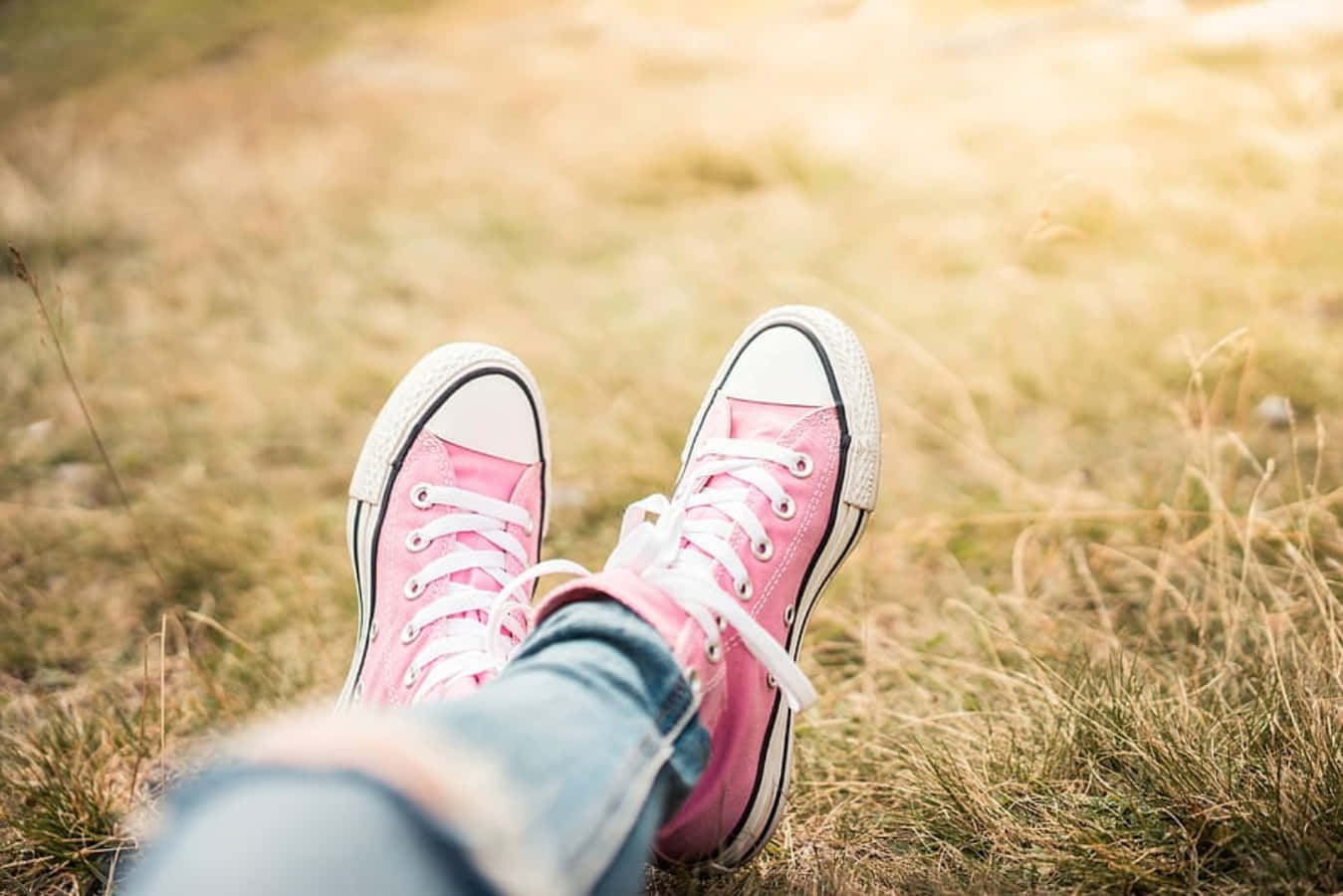 Pink Shoes 桌布 壁紙 — 粉紅色運動鞋配白色鞋帶擱在金黃色的草地上，雙腿在陽光明媚的田野中放鬆地晃動。