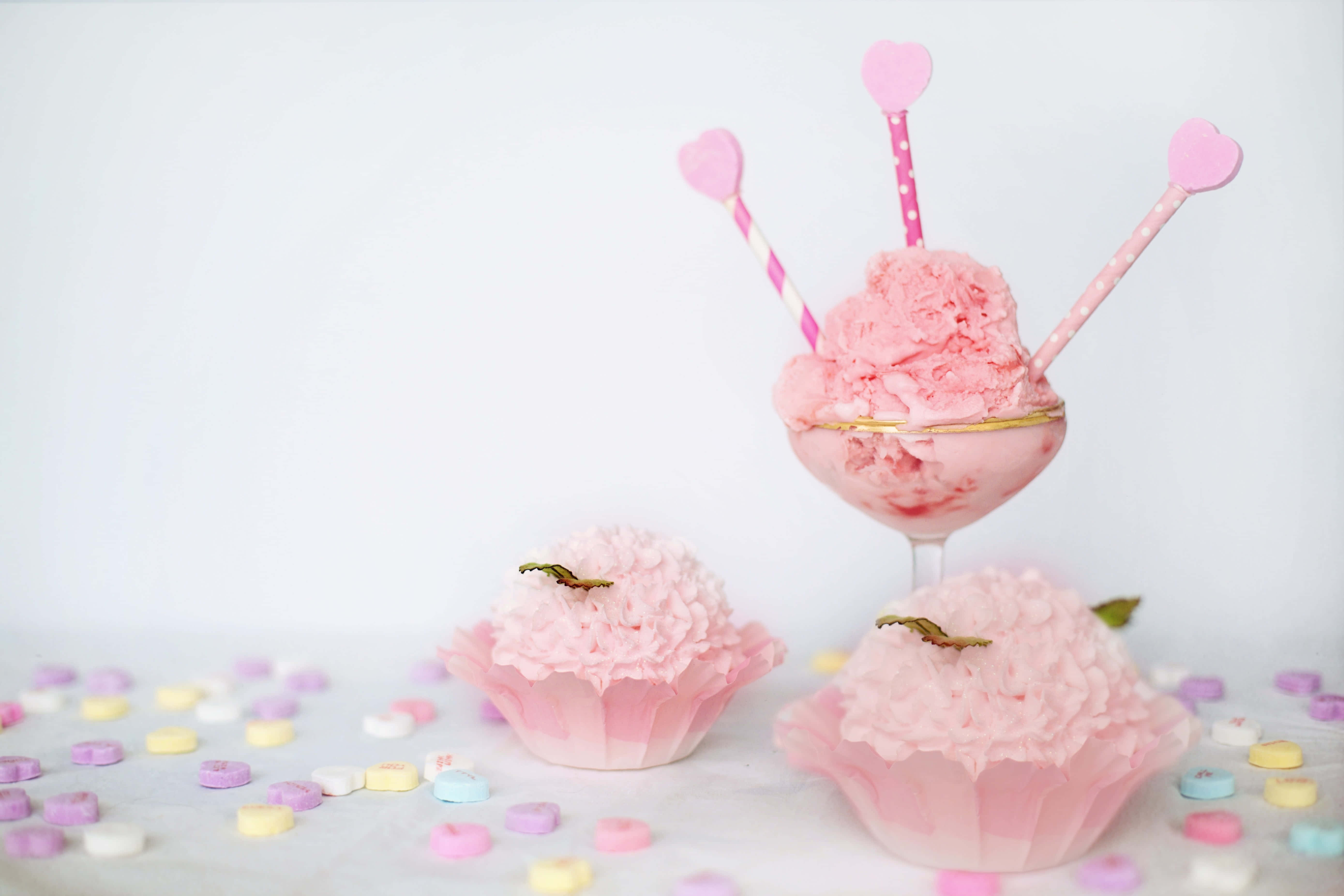 Pink Cupcakes 桌布 壁紙 — 粉紅色雪糕放在玻璃杯內，配上心形飲管和杯子蛋糕，周圍散落著色彩繽紛的心形糖果，置於白色表面上。