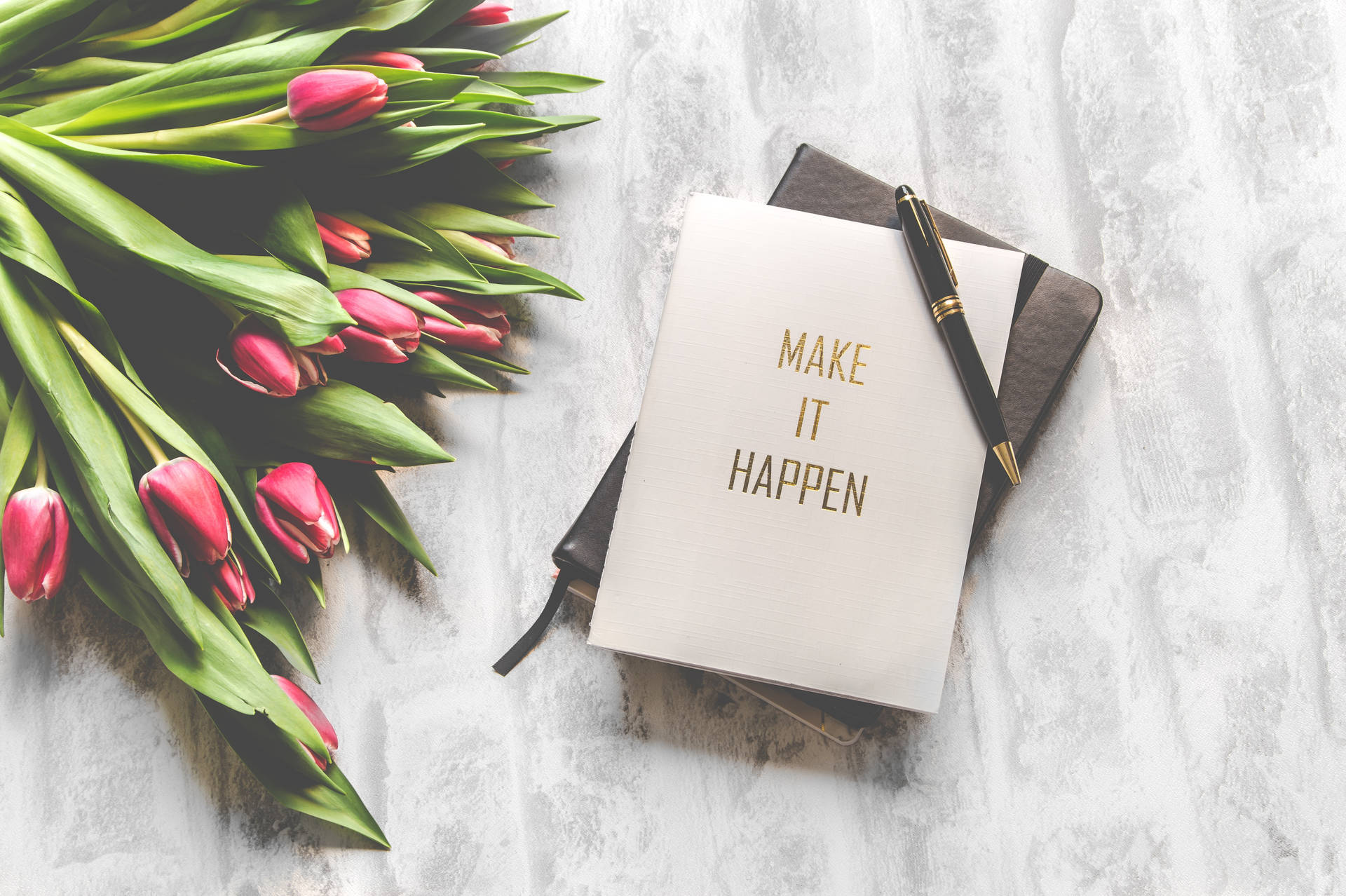 粉紅色鬱金香和一本帶有「MAKE IT HAPPEN」文字的筆記本，放在白色有紋理的表面上。
