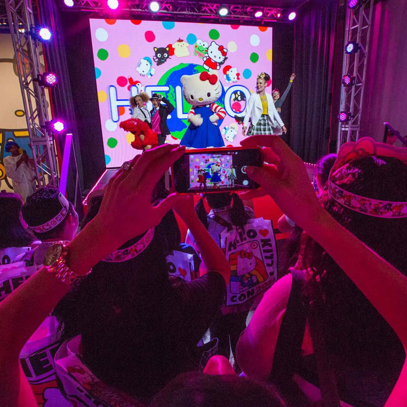 Kawaii Hello Kitty 桌布 壁紙 — 粉絲們在熱鬧的展會上，拍攝站在充滿活力的粉紅色燈光舞台上的Hello Kitty吉祥物。