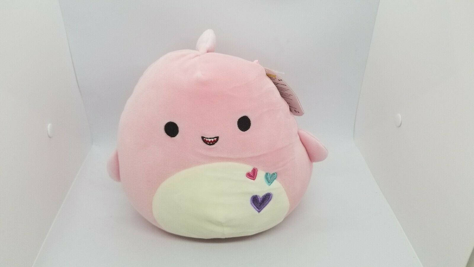 粉色Squishmallows玩偶盒中 壁紙 — 一個粉紅色毛絨玩具，面帶微笑，身上有彩色心形圖案，放在一個白色盒子裡。